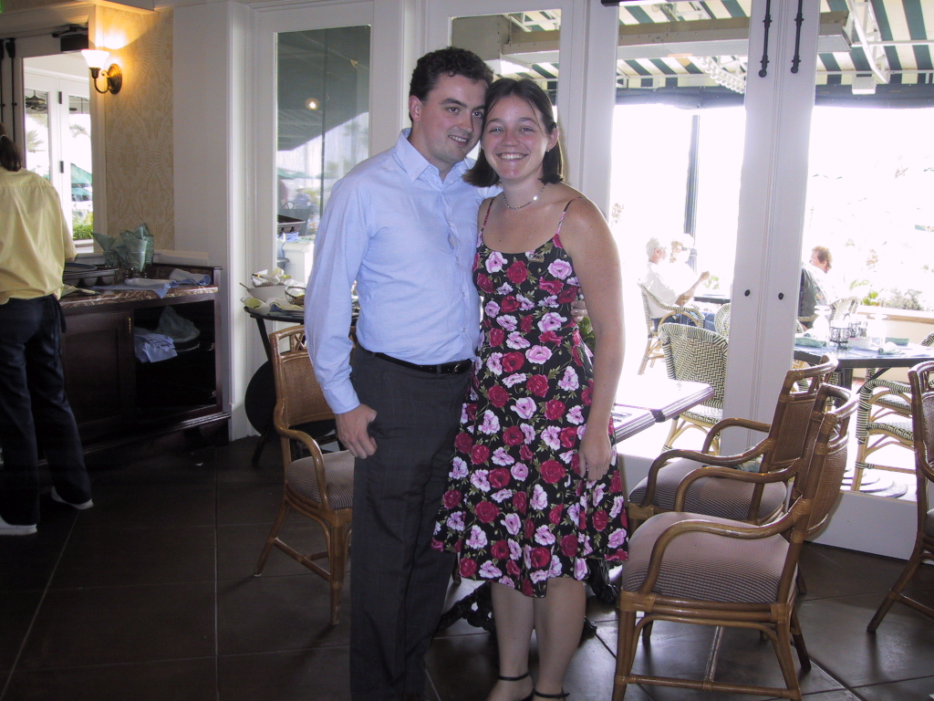 Two people pose for a photo while standing in front of a table in a restaurant. The person on the left is wearing a blue shirt and gray pants. The person on the right is wearing a red and pink floral dress. They’re both smiling.