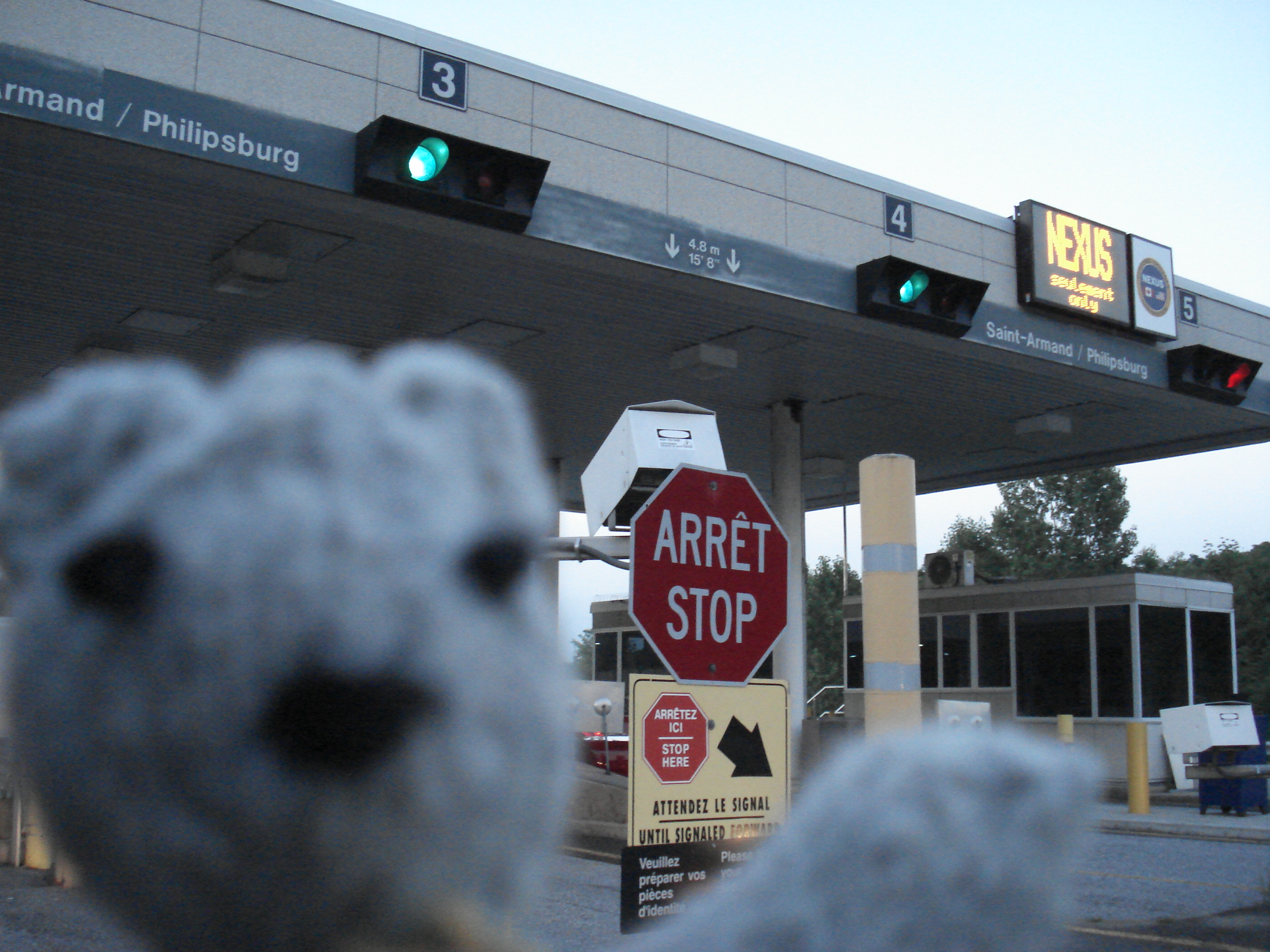 An out-of-focus toy bear knit of gray wool appears before an in-focus US/Canada border crossing. All of the sinage is in French and English, including a red octagonal stop sign labeled both ARRÊT and STOP.