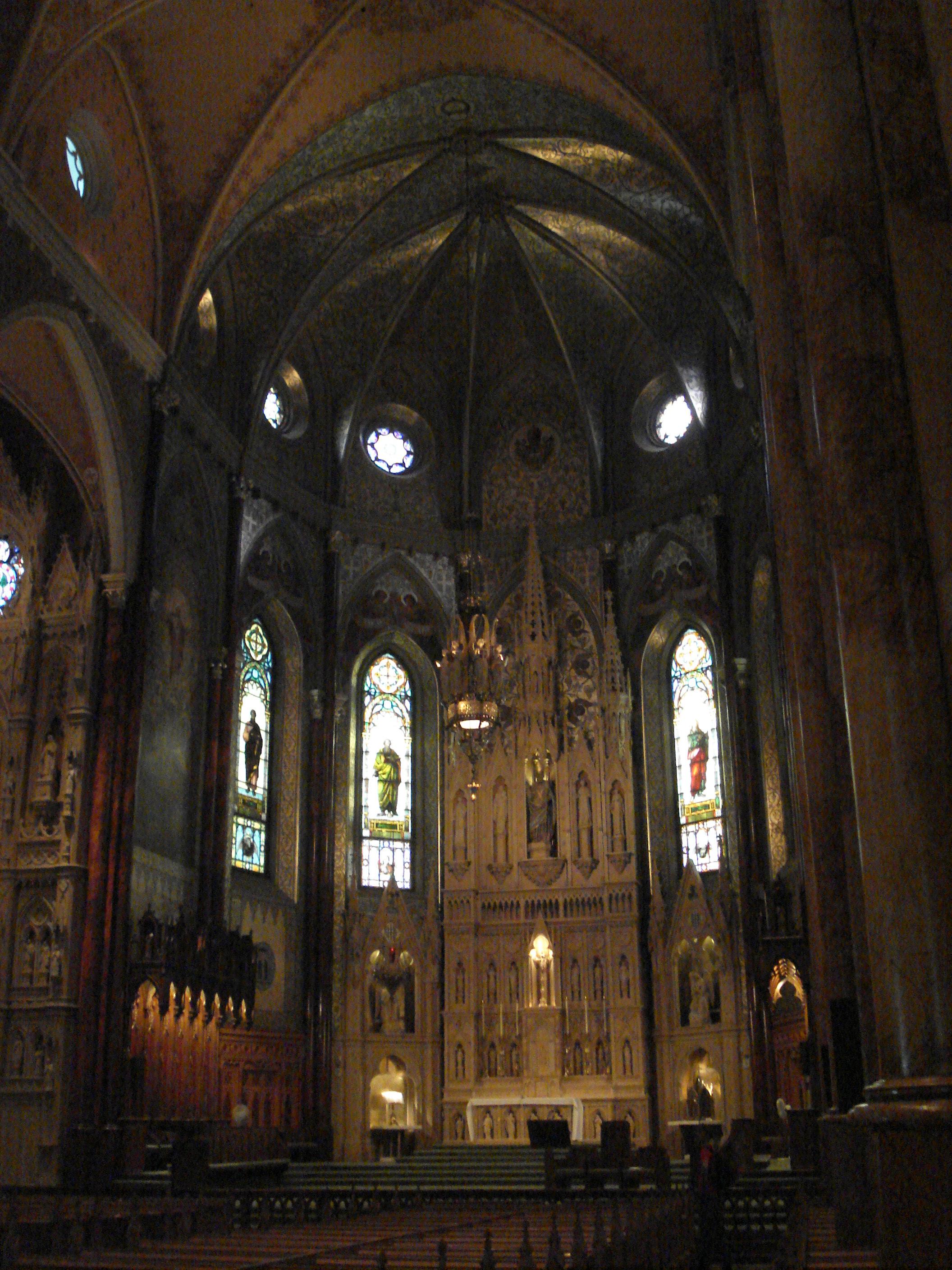 The dark interior of a large Catholic church is photographed in portrait, which gives the viewer a sense of just how tall the interior must be (which is very tall). Various stained glass windows are visible, as are a number of pews.