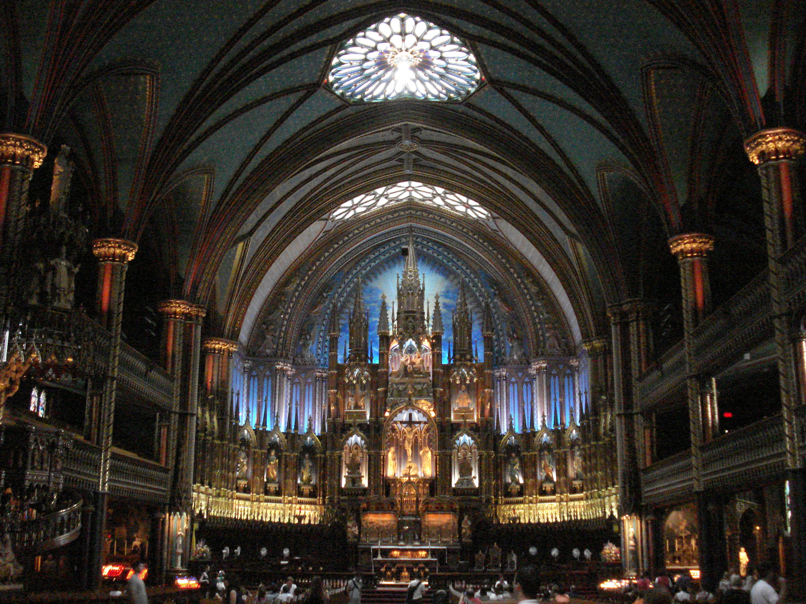 A photograph of the interior of a large, ornate Catholic church. Stained glass in the ceiling helps to light the room. The elaborate altar backdrop has many statues and a number of blue elements which draw the eye.
