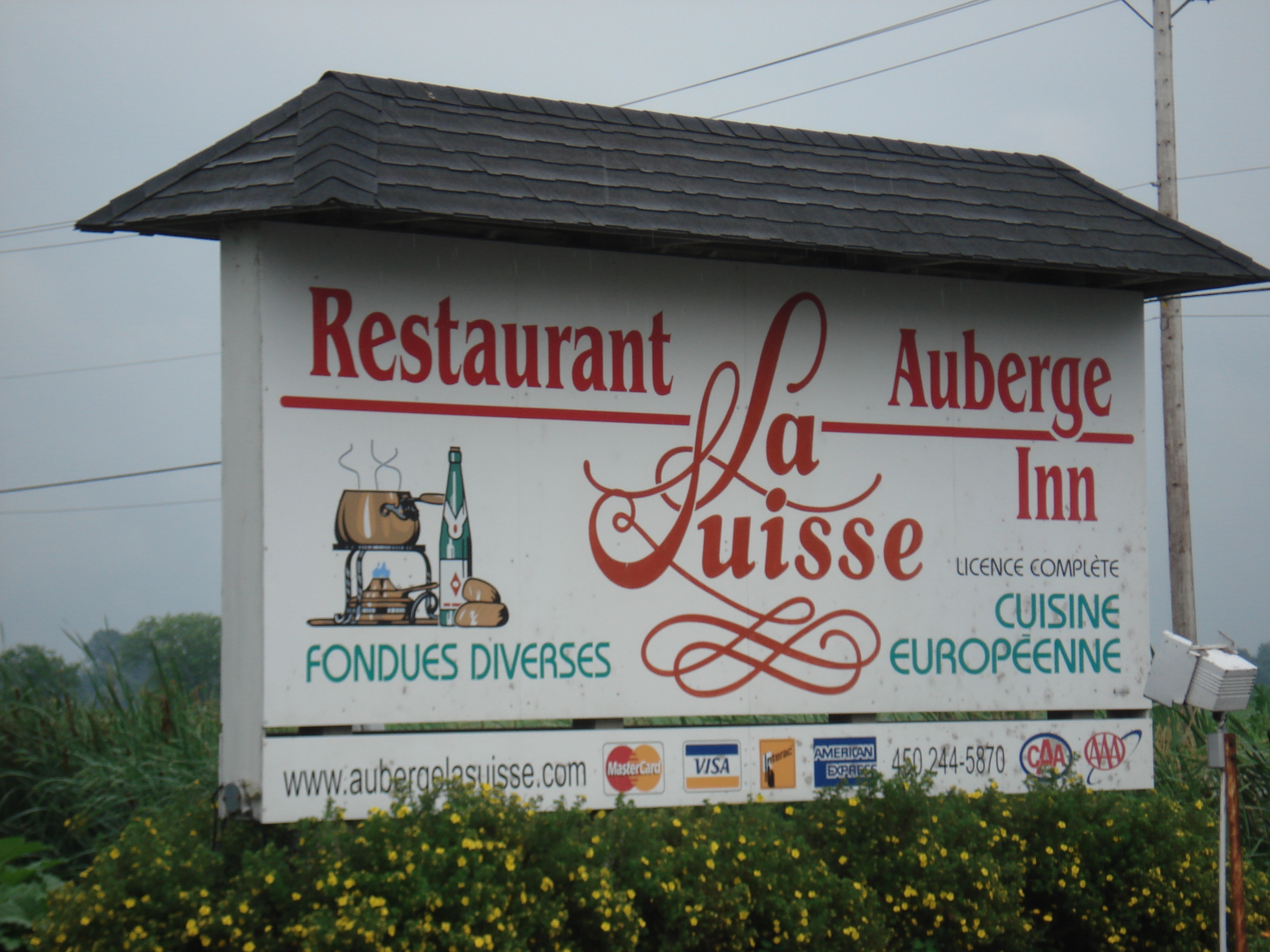 A French-language sign on the side of a road advertises a bed and breakfast called Auberge La Suisse. It says their restaurant serves fondue of many kinds as well as other European cuisine.