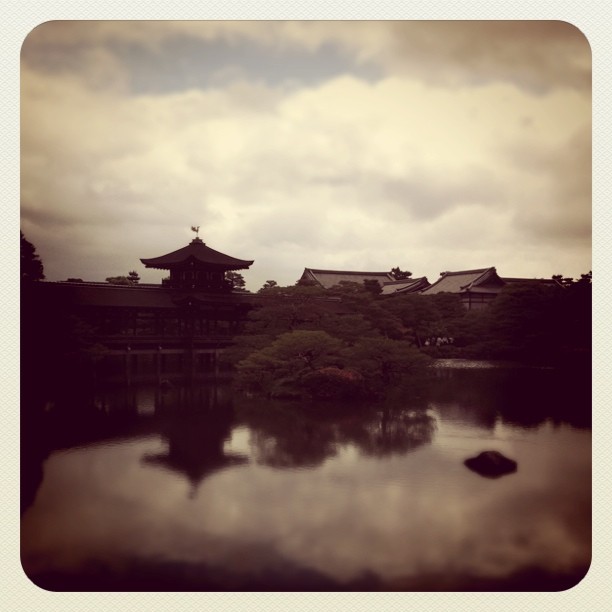 Several dark buildings in traditional Japanese architecture sit just on the other side of a small lake or pond. A cloudy sky hangs above them.