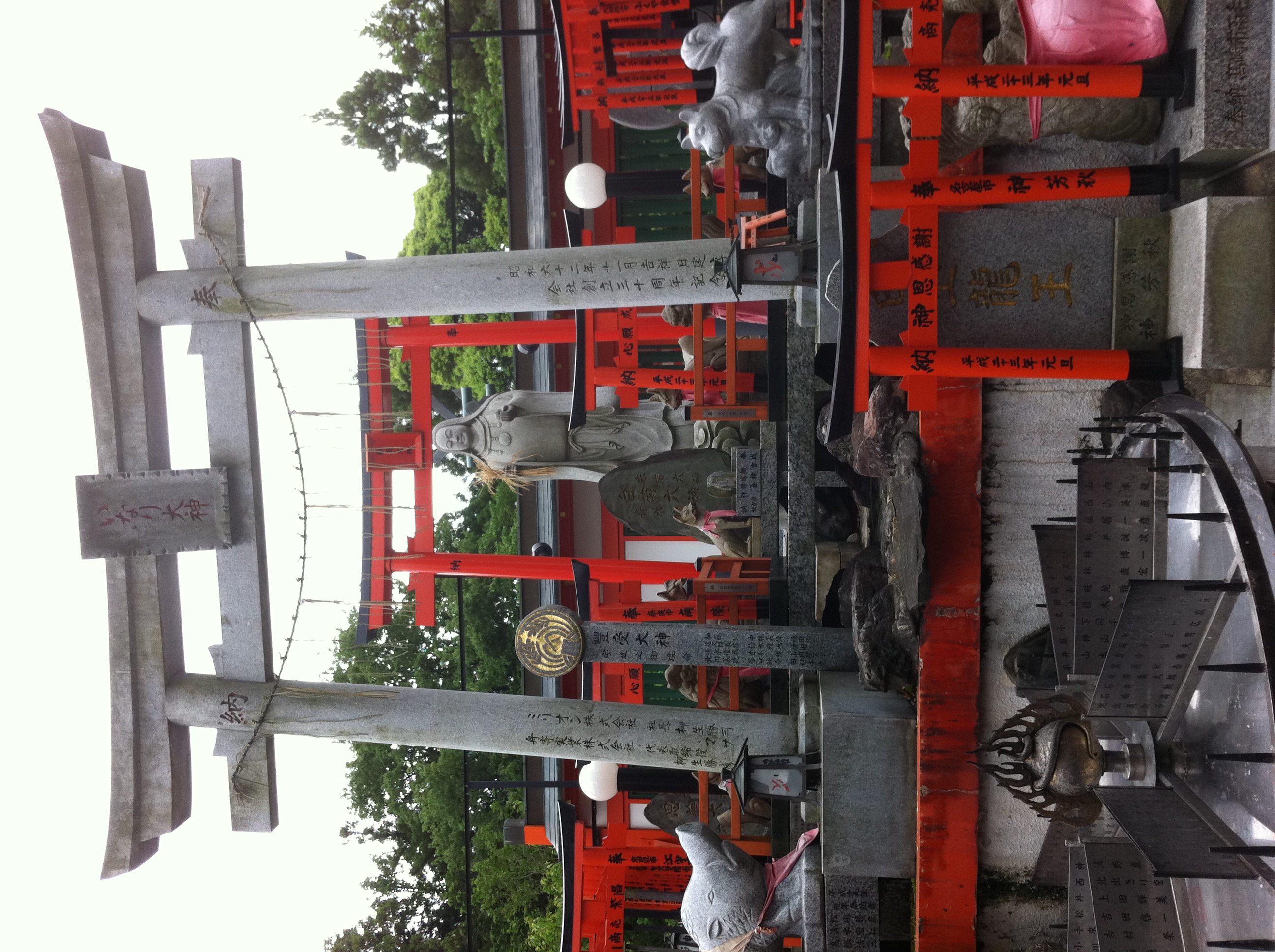Many small vermillion torii gates and kitsune statues are scattered under a larger, stone torii. Beyond the stone torii, a tall statue of Inari-okami is visible.