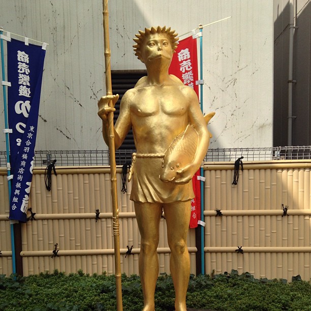 A gold statue of a somewhat fishy-looking fellow (in the literal ’like a fish’, sense, not in the ’shady’ sense) holding a fish in his left hand and a bamboo staff in his right.