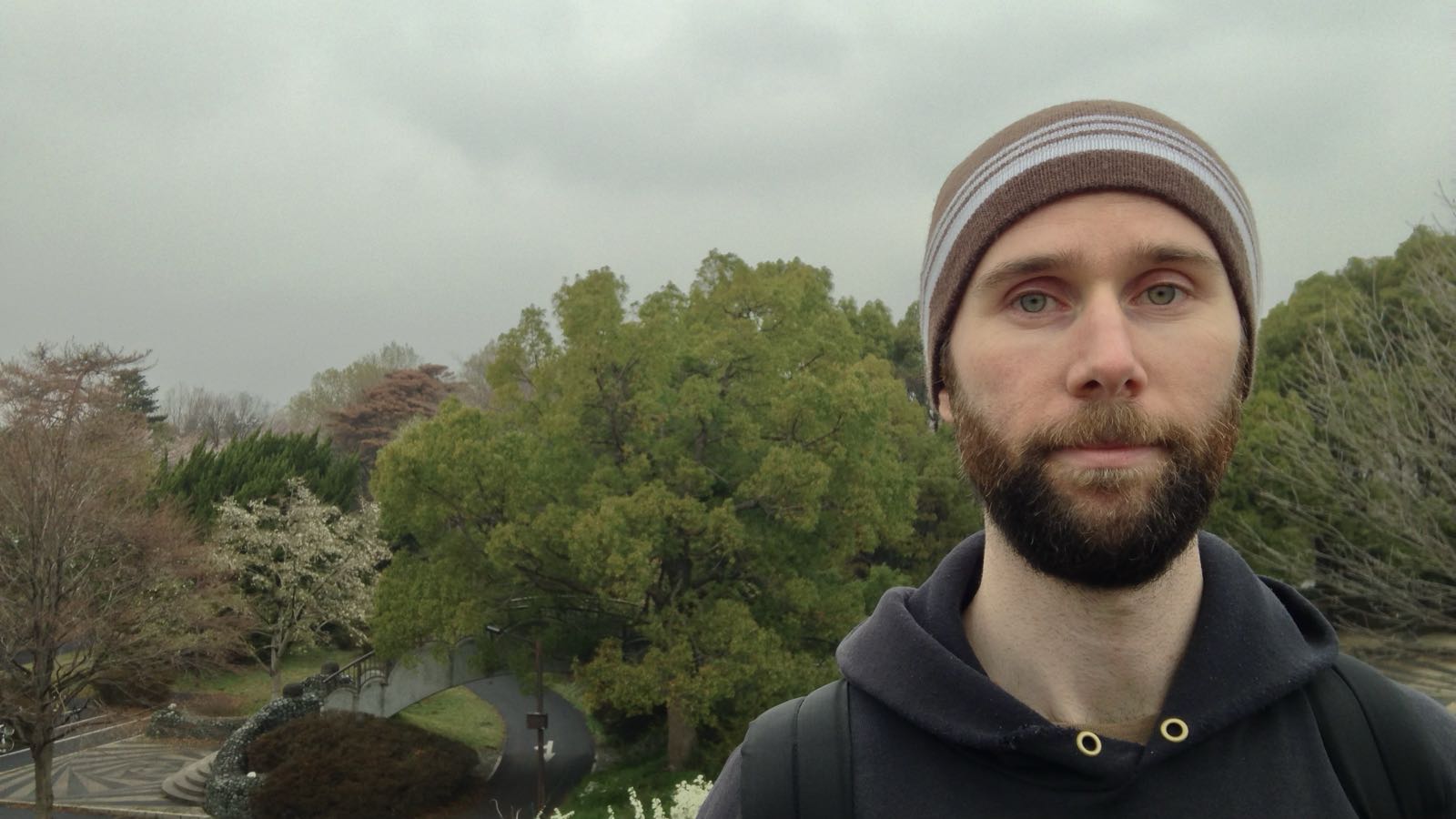 A man with a neutral expression faces the camera. He’s wearing a hoodie and a beanie, and has facial hair. Behind him, trees with autumnal leaves are visible.