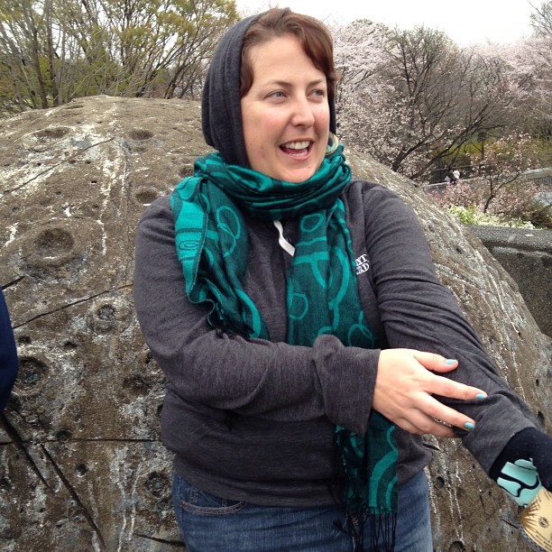 Melissa wears a green scarf over a gray hoodie. She’ standing in front of a stone that has moon-like crater in it.