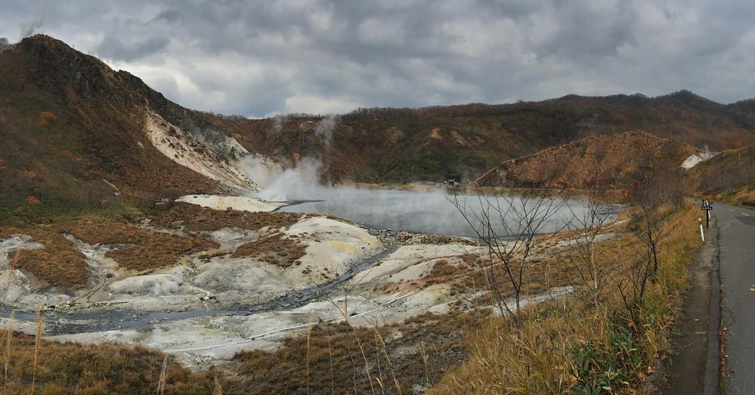 Mountainous, predominantly brown terrain. In the center of the frame is a lake, with a whole lot of steam coming off of it. A road is visible in the right of the frame.
