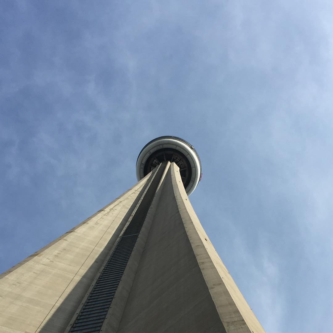 The camera looks up toward the sky from the base of a large, concrete tower. There’s a circular structure at the top of the tower, in the center of the frame. There’s a partly cloudy sky beyond the tower.