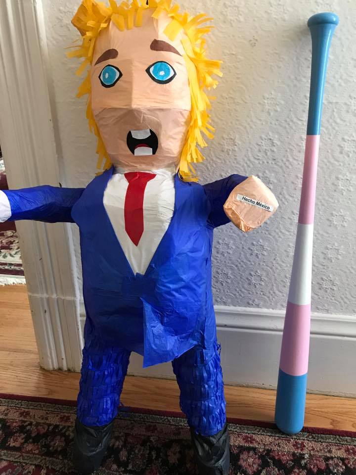 A piñata of Donald Trump leans against a wall. Just to its right, a baseball bat painted with the transgender rights flag also leans against the wall.