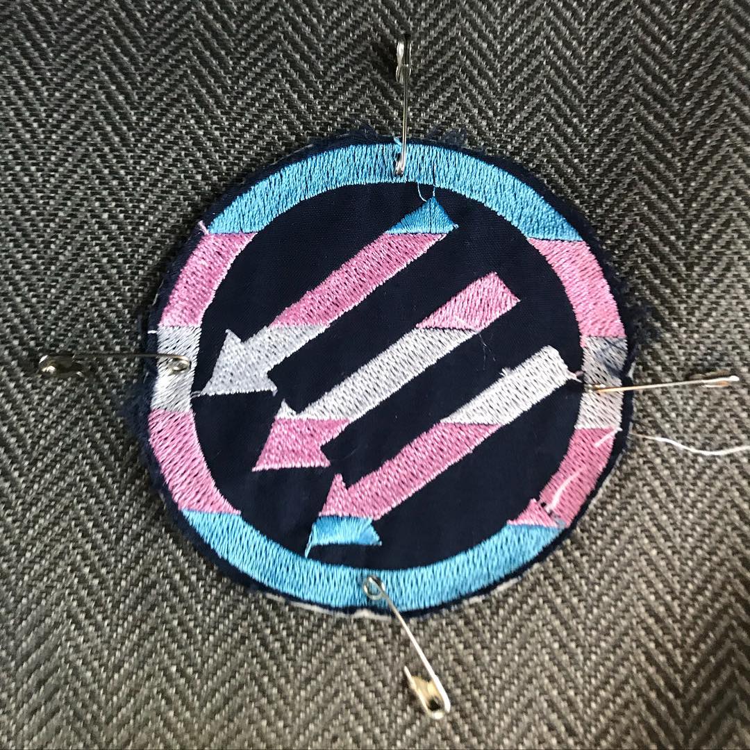 Some gray herringbone fabric has a cloth patch attached to it with safety pins. The patch depicts the three arrows of the Iron Front, an antifascist group active in Germany during the Third Reich. The arrows are rendered in the colors of the transgender pride flag.