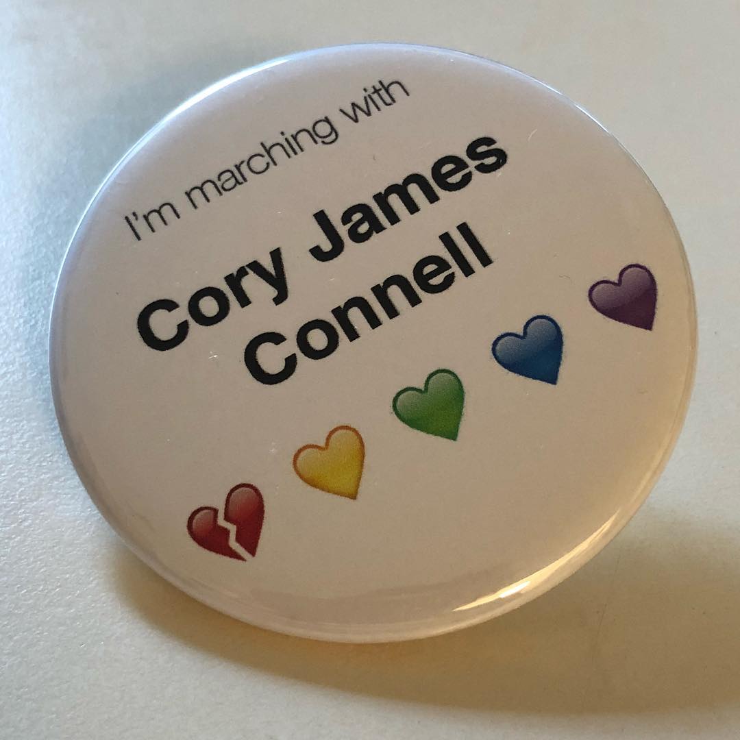 U+1F9E1 ORANGE HEART (🧡) had yet to be added to Unicode—hence its absence on the pin. A large, white pin rests on a white surface. The pin reads “I’m marching with Cory James Connell 💔💛💚💙💜”.