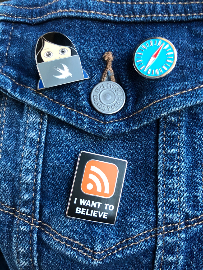 a pocket of a denim jacket. there are three enamel pins on it: one of a woman Swift developer, one of the Safari icon, and one of the “I want to believe” poster from the x-files, but with an RSS logo instead of the UFO.
