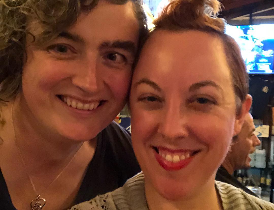 Another close-up of two smiling women. The one on the left has curly hair and strabismus. The one on the right has this one curly tuft front and center in her ginger hair.