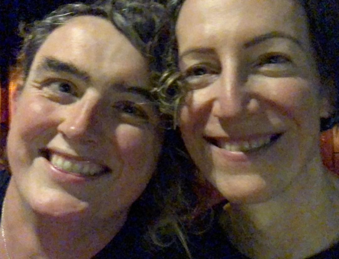 Two people smile at the camera. The photo was taken in low light, so it’s a bit grainy. The one on the left has curly hair.