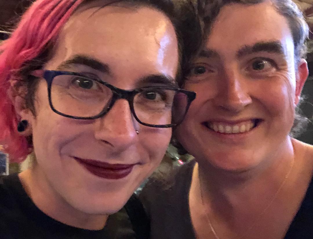 A close-up of two people’s faces. The one on the left has pink hair and wears glasses.
