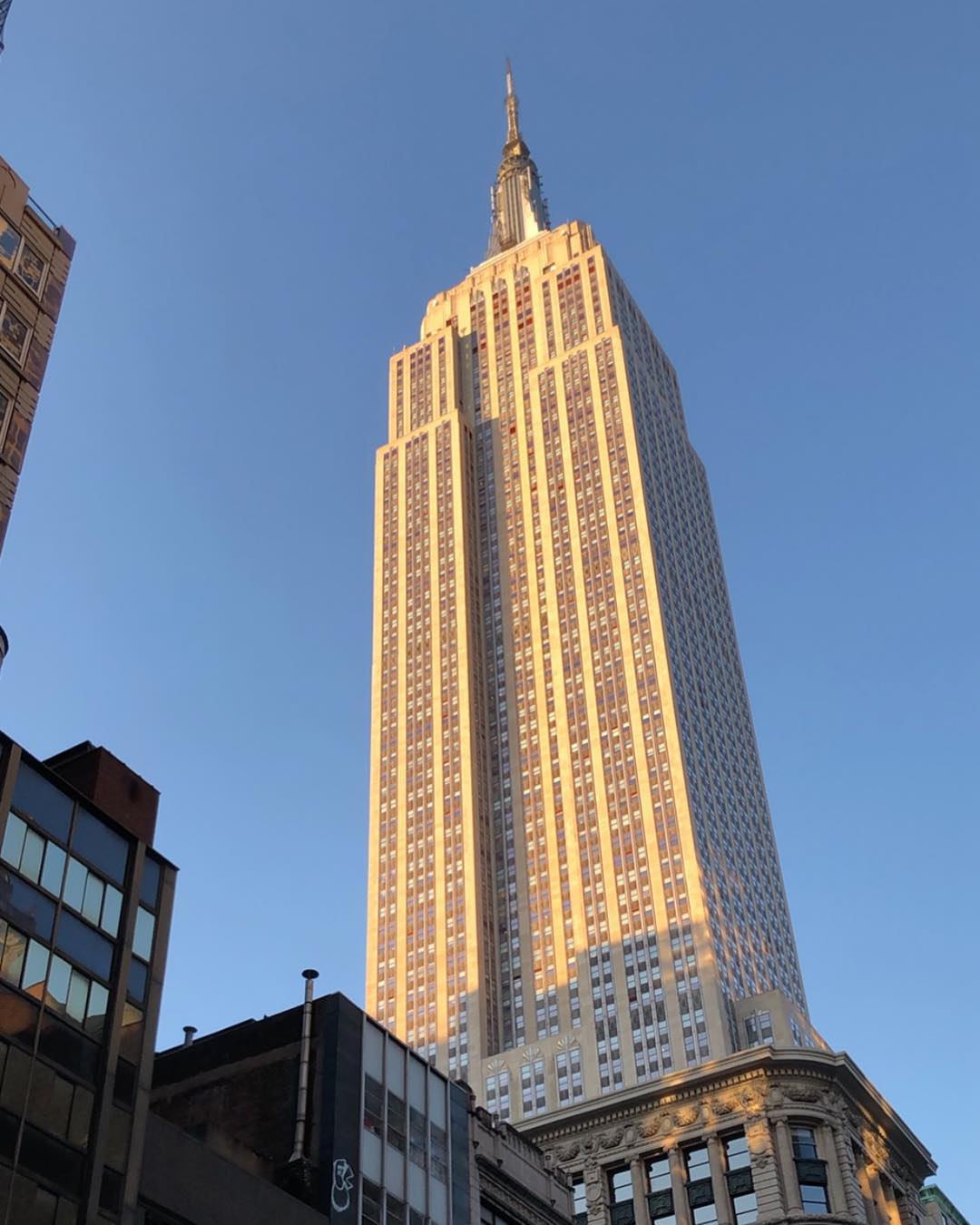 The Empire State Building, bathed in sunlight, reaches for a clear sky above Manhattan. The photo is at something of a jaunty angle, and while there are some other buildings visible around the bottom and left edges, the frame is completely dominated by the old skyscraper.