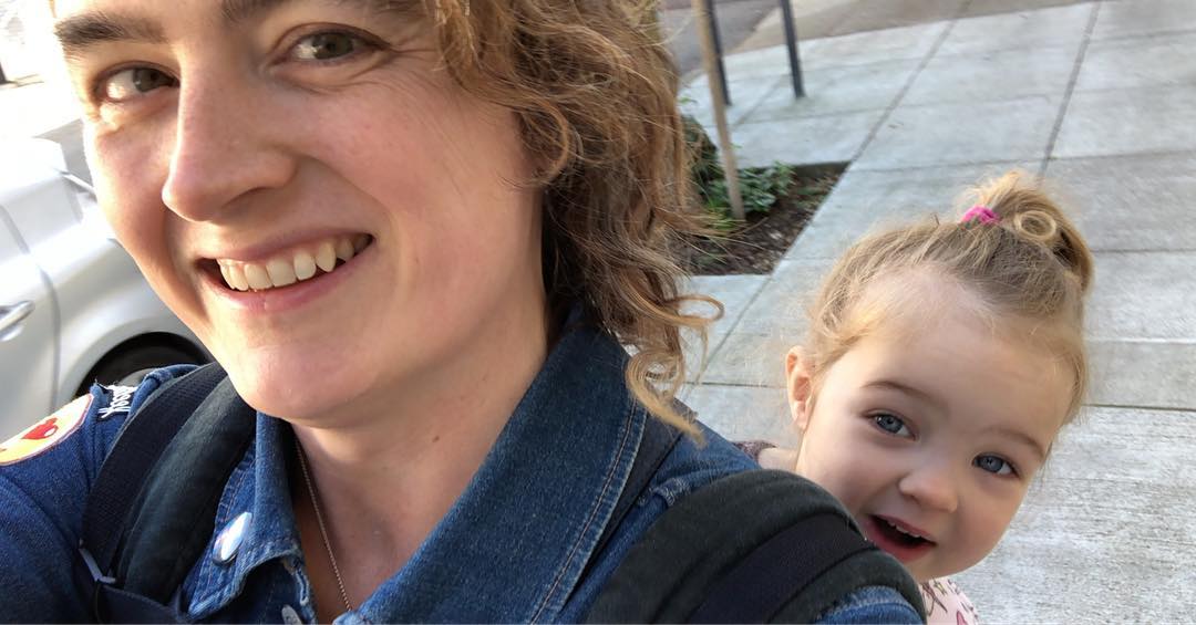 A selfie of a woman wearing a toddler on her back.