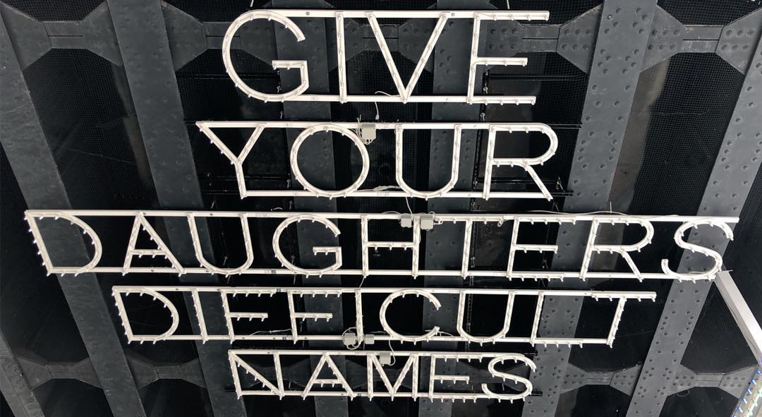 A metal, lit sign hangs from some kind of metal structure. The sign reads “GIVE YOUR DAUGHTERS DIFFICULT NAMES”.