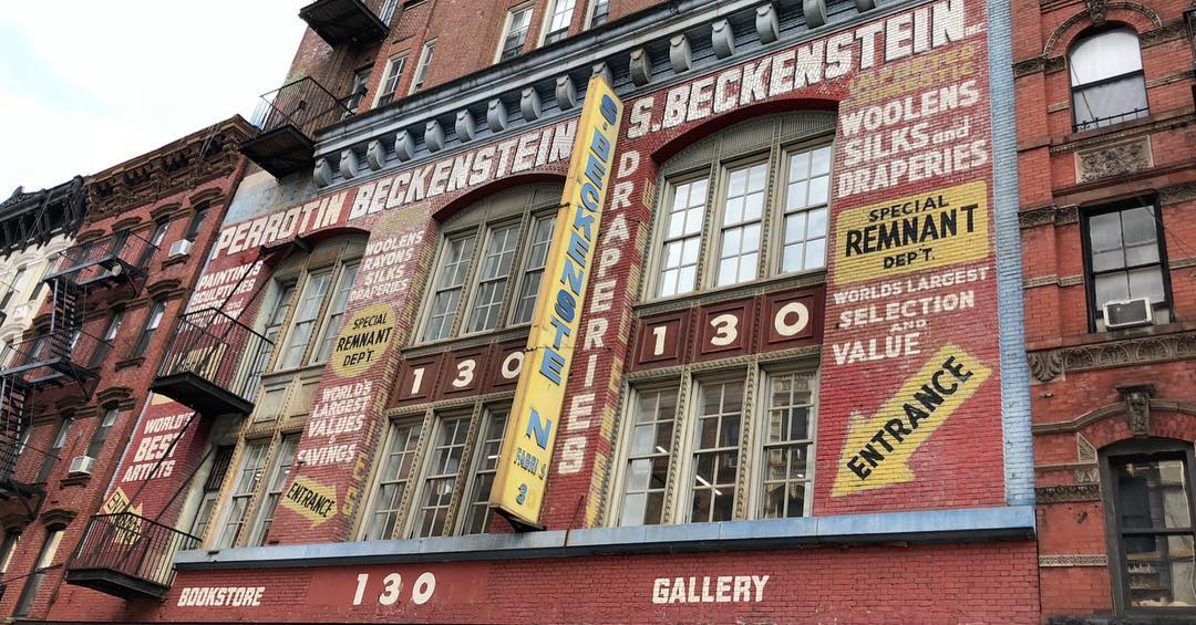 An old brick warehouse in Manhattan is covered in old-timey painted signs advertising its business. They read: “PERROTIN BECKENSTEIN”, “PAINTINGS, SCULPTURES”, “WOOLENS, RAYONS, SILKS, DRAPERIES”, “SPECIAL REMNANT DEP’T”, “WORLD’S BEST ARTISTS”, and the like.
