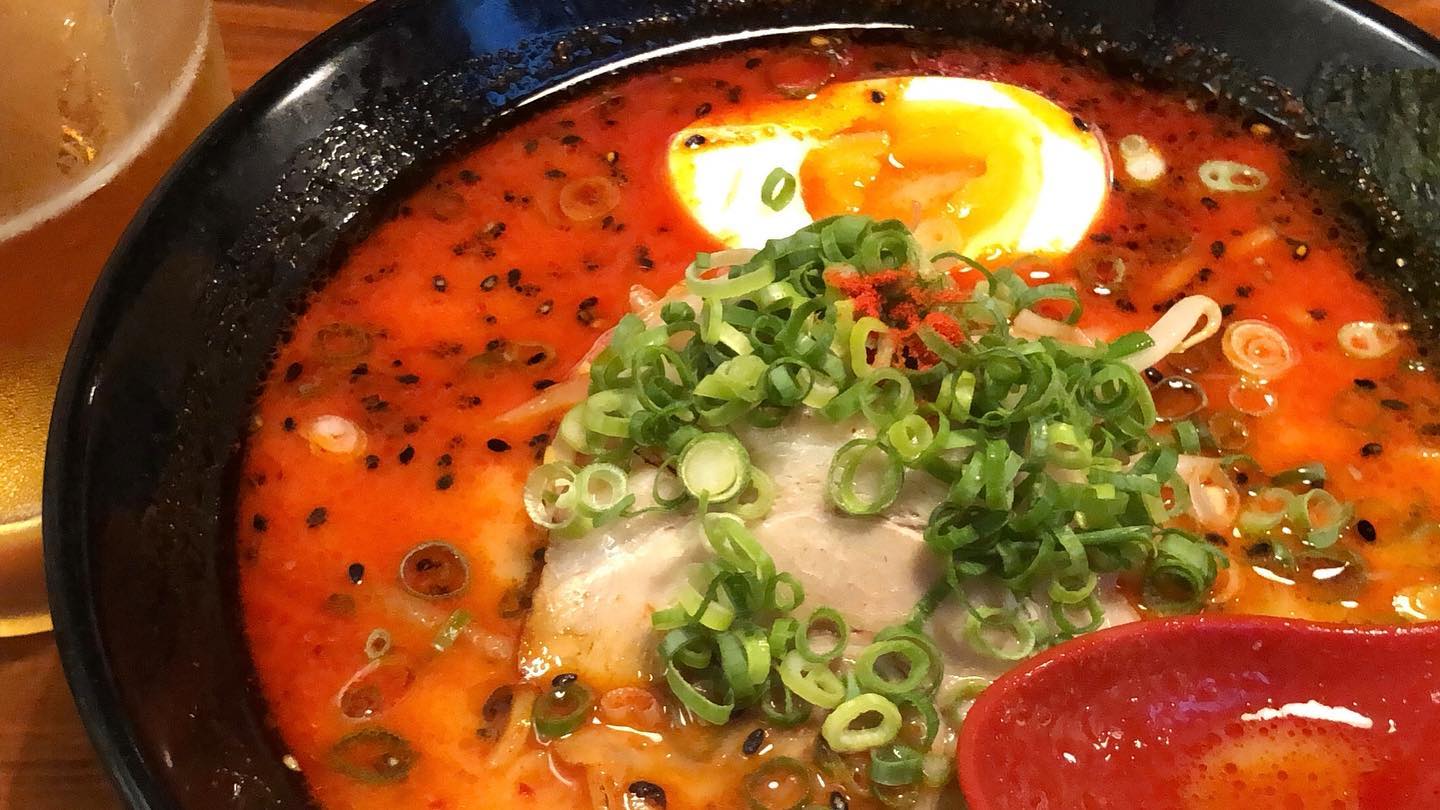 A bowl of ramen. The broth is quite red, suggesting it’s quite spicy. There’s a whole pile of green onion on top of pork chashu in the bowl. An egg is visible at the top edge of the bowl.