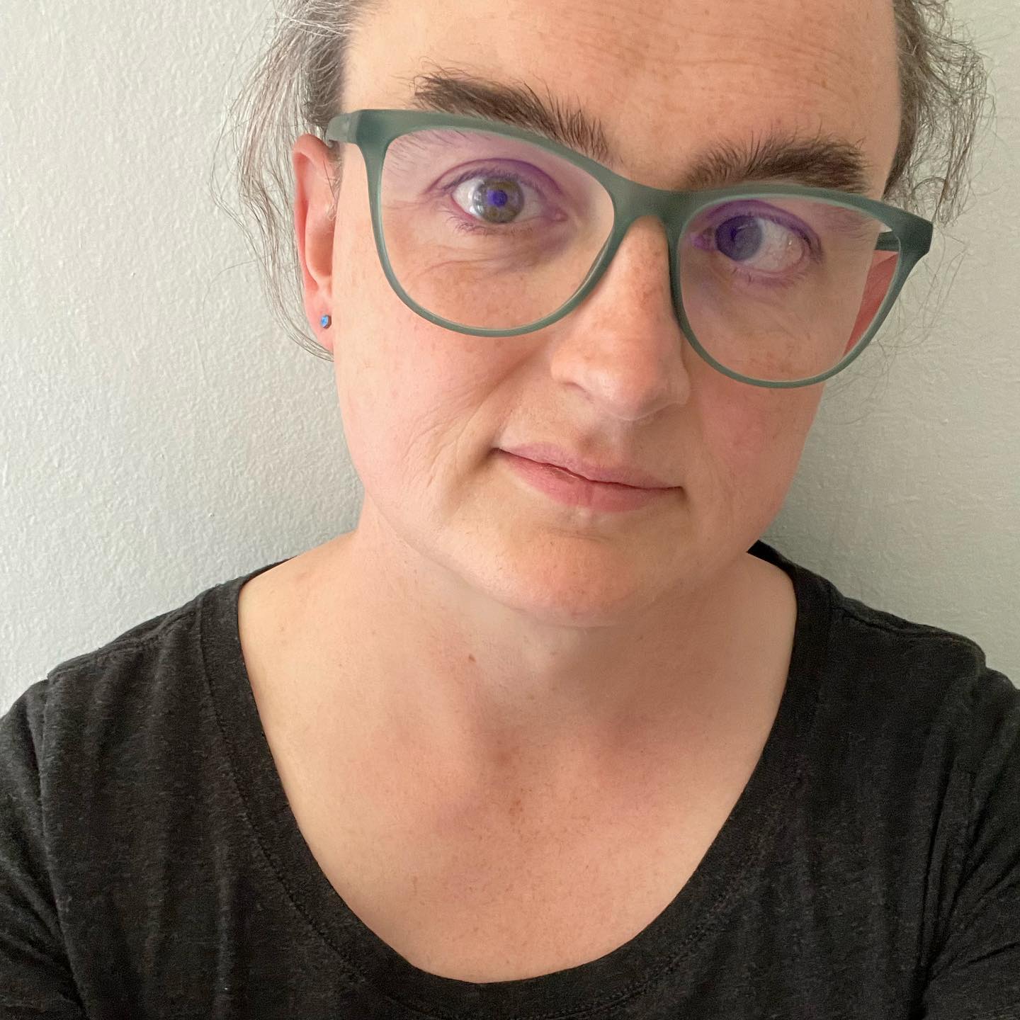 A woman hazel eyes looks at the camera. She’s wearing a black T-shirt with a scooped neckline, and glasses with a grayish-blue frame. She has an ambivalent expression on her face.