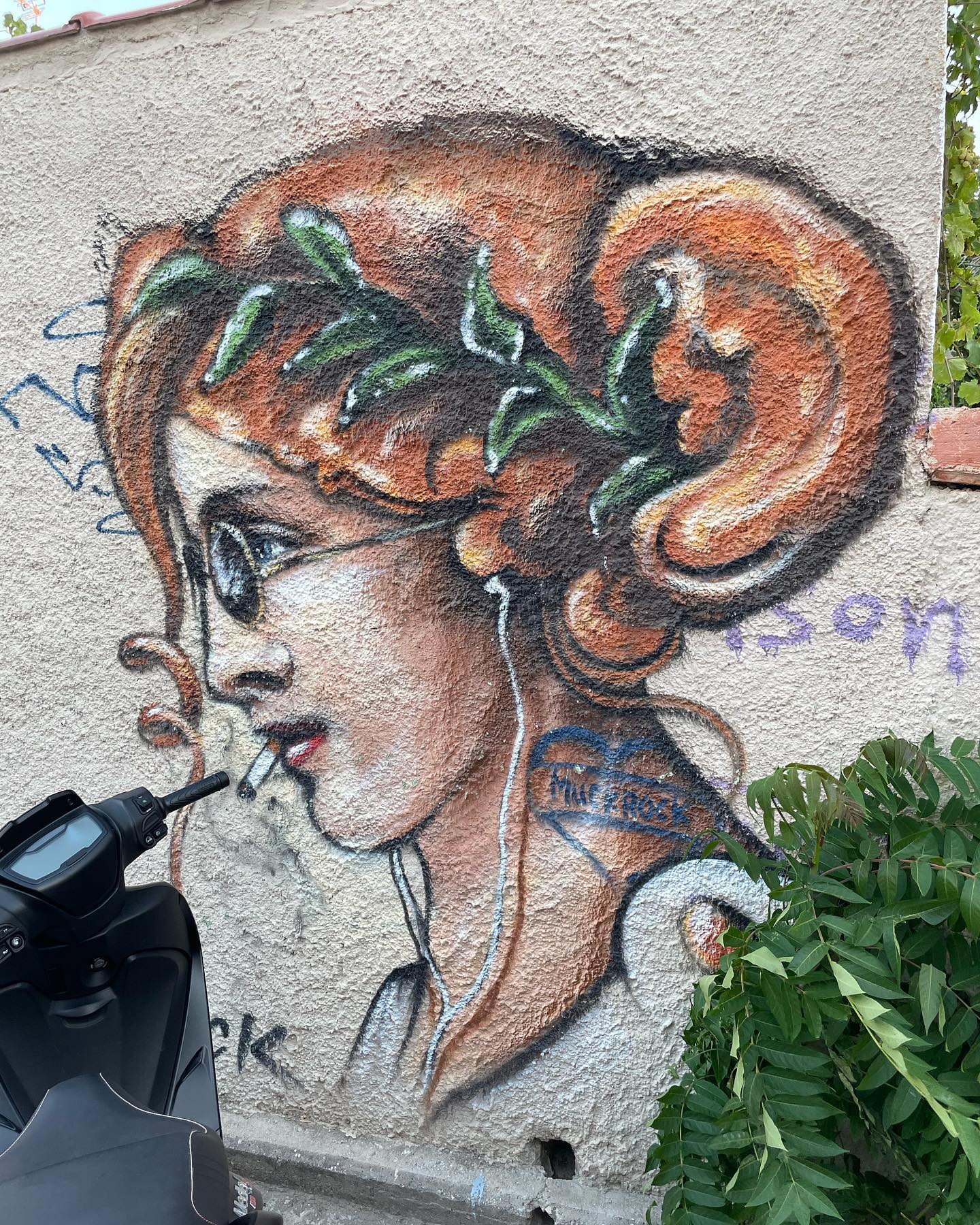 Street art of a red-headed woman. The painting is on the wall of
  a beige, stucco building. The woman is wearing round, dark sunglasses,
  a crown of laurel leaves, and white clothing. She’s smoking a
  cigarette and has white headphones in her ears (Apple EarPods,
  presumably). Her long hair is up, but some of it has escaped and hangs
  in curls.