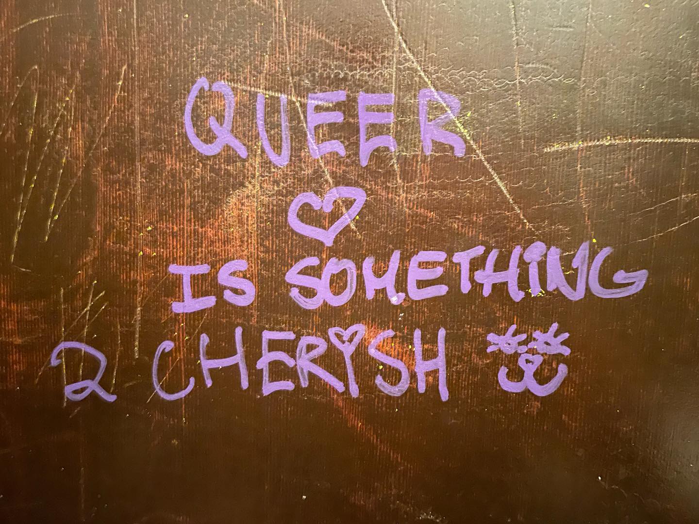 On a brown wooden wall, “QUEER ❤ IS SOMETHING 2 CHERISH” is written in lavendar ink.