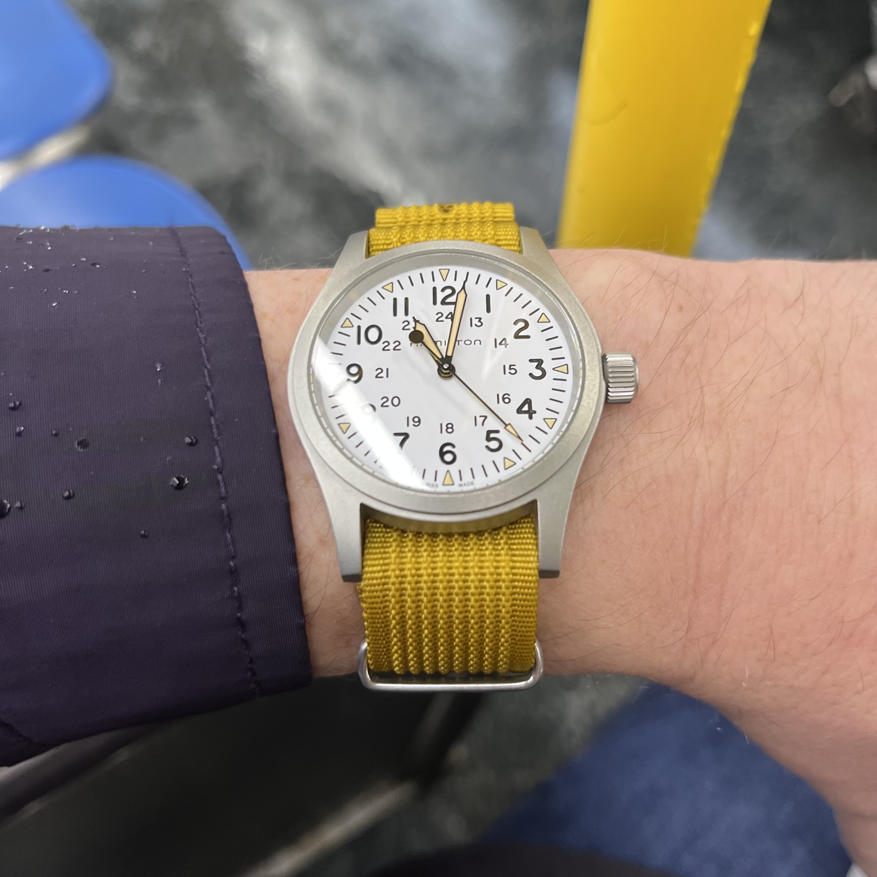 A wrist shot of a white dial field watch on a mustard yellow NATO strap.