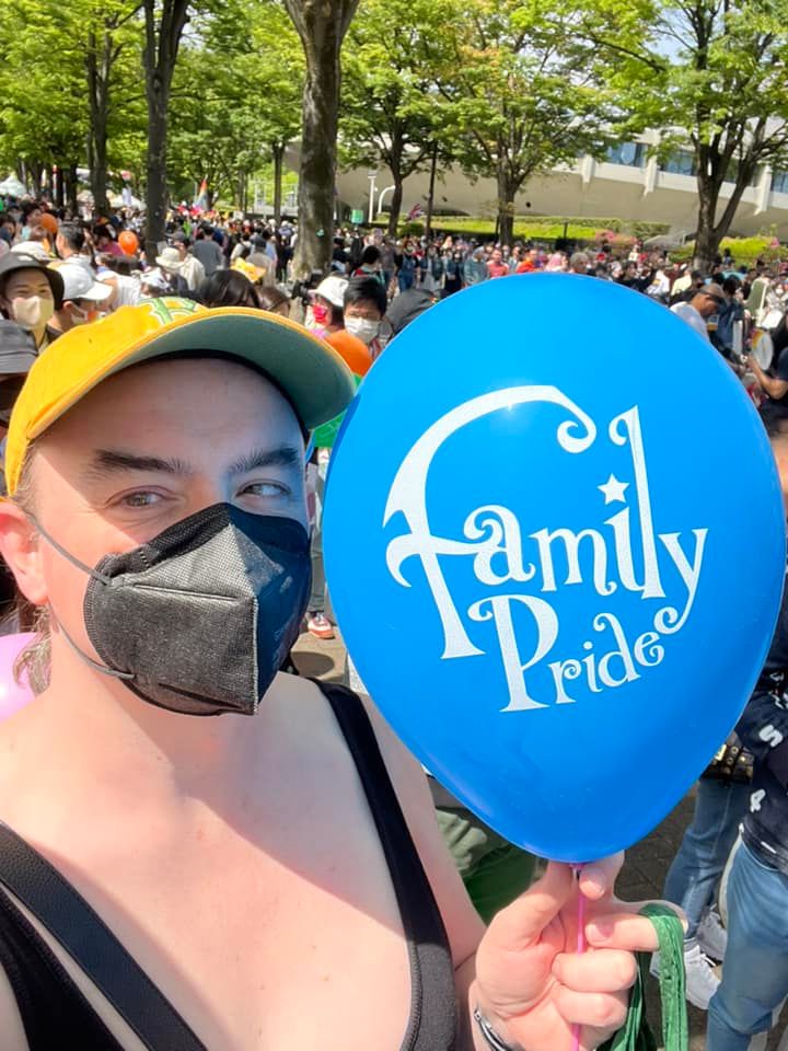 A girl in a yellow baseball hat and a black kN95 mask holds a blue balloon with the words “Family Pride” printed on it. Behind her, a large crowd of people is visible.