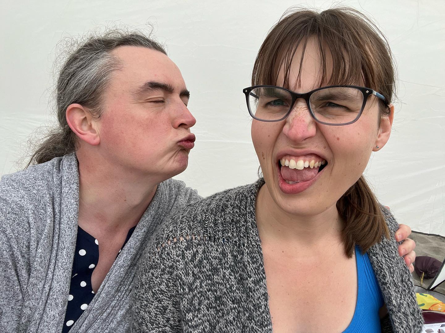 I am trying to kiss Eryn in a very goofy way.
She is not having it; her facial expression is one of mock-disgust.
We’re both wearing gray cardigans.
My hair is clearly more gray than I care to admit.