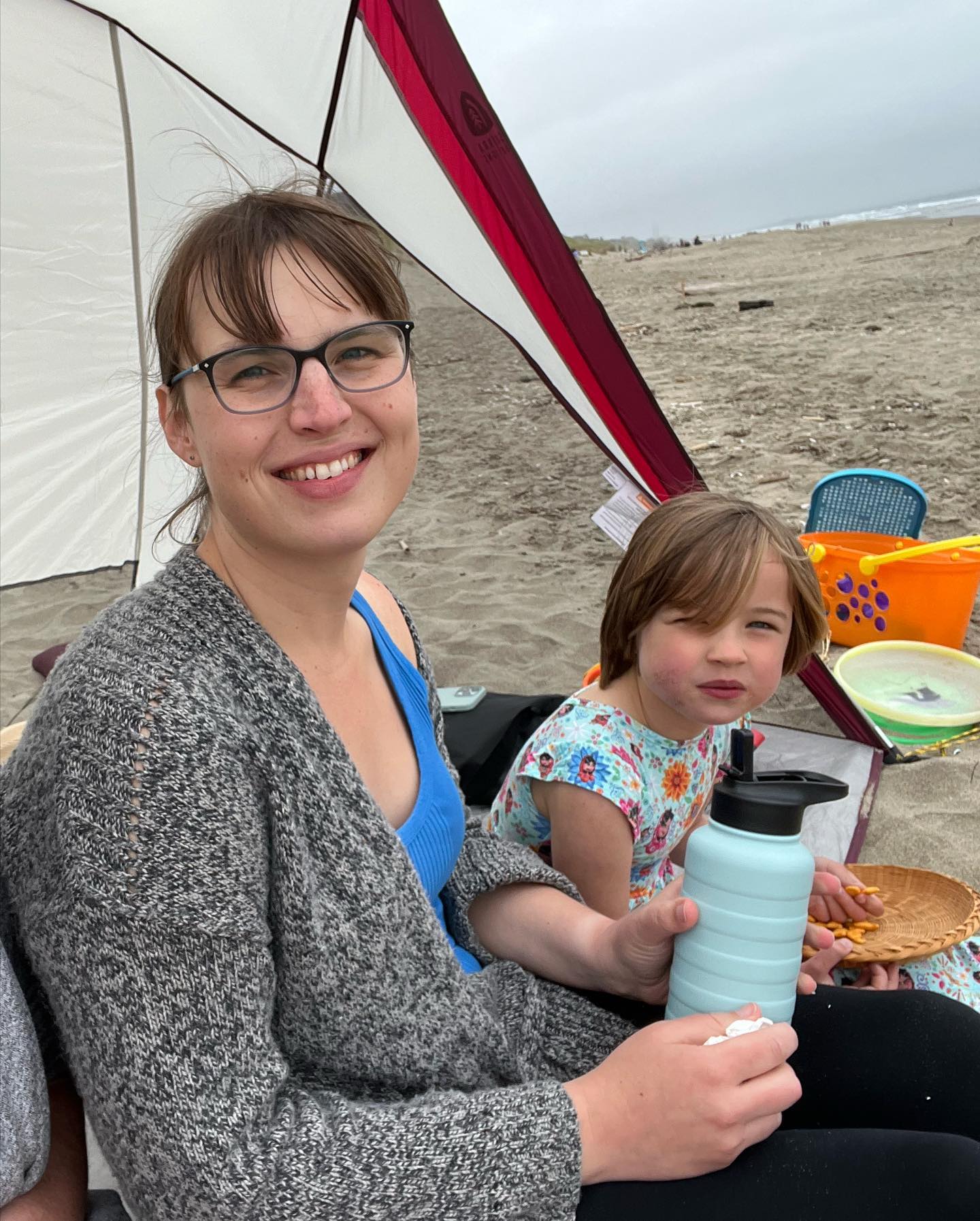 Eryn and EJ sit in a half-tent beach shelter thingy on the beach.
Eryn is smiling at the camera, while EJ glares at it.
Some of EJ’s beach toys are visible on the sand behind her.
Eryn’s holding a water bottle.
The weather doesn’t seem super great for a beach day, but it didn’t stop us.