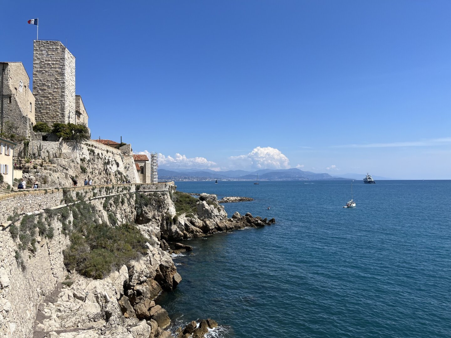 Overlooking the Mediterranean, an off-white Medieval stone tower stands tall, with the Tricolour of France flying from a flagpole at the top.