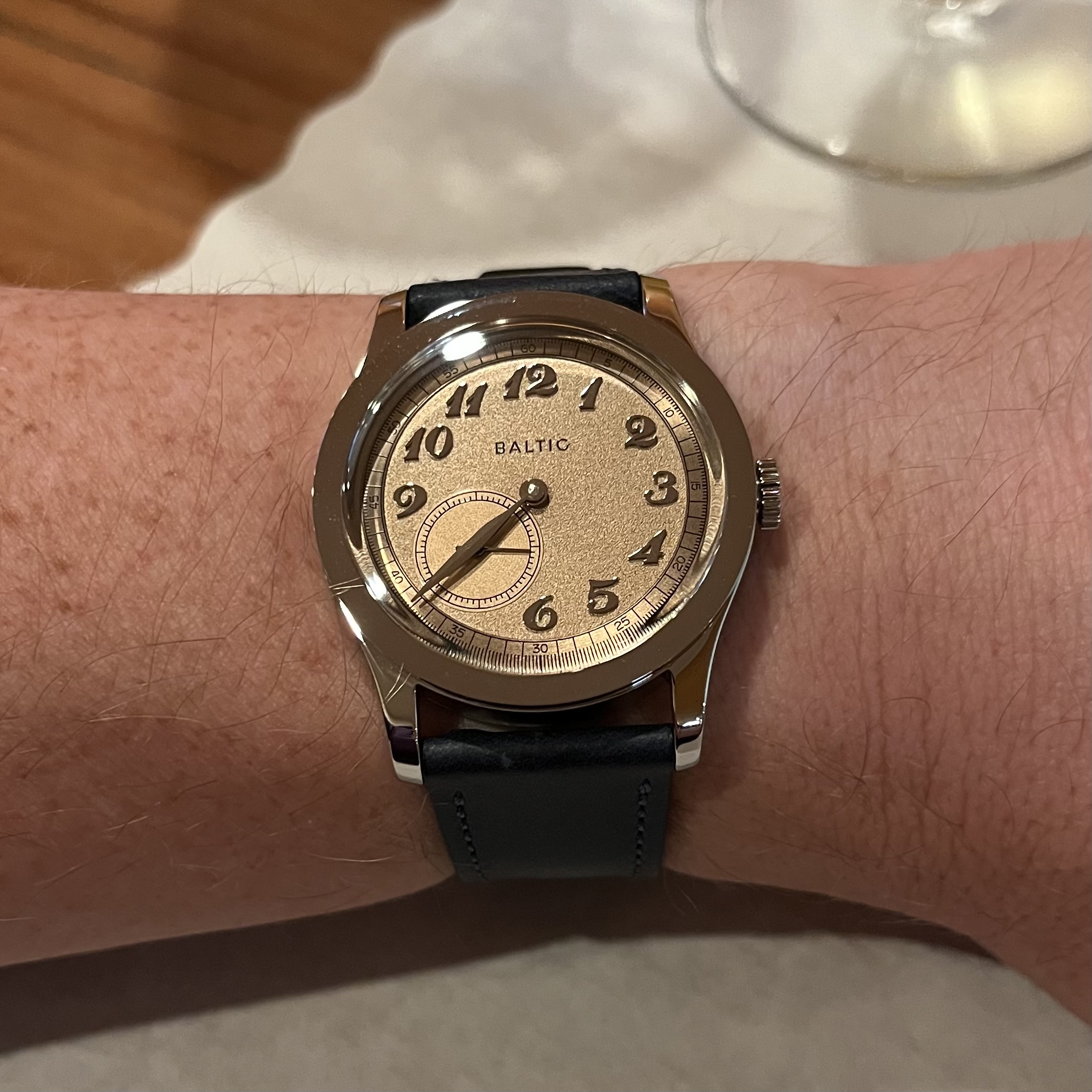 a freckled wrist is wearing a shiny dress watch. The watch's case and lugs are polished stainless steel. Its dial is salmon-colored, with applied Breguet numerals and a small seconds subdial in the bottom left. The main section of the dial has a grainy texture, whereas the subdial has a radial finish.