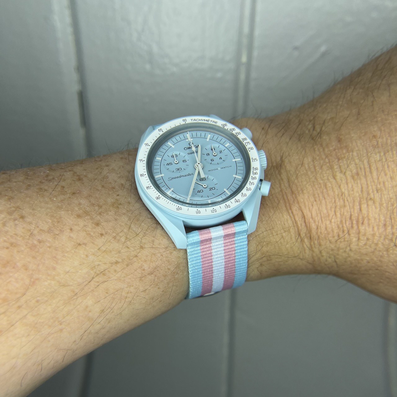 The wrist of a woman with a watch on it. The watch is an Omega × Swatch Bioceramic Moonswatch (specifically, the baby-blue colored Mission to Uranus). It's on a nylon strap in the colors of the trans pride flag.