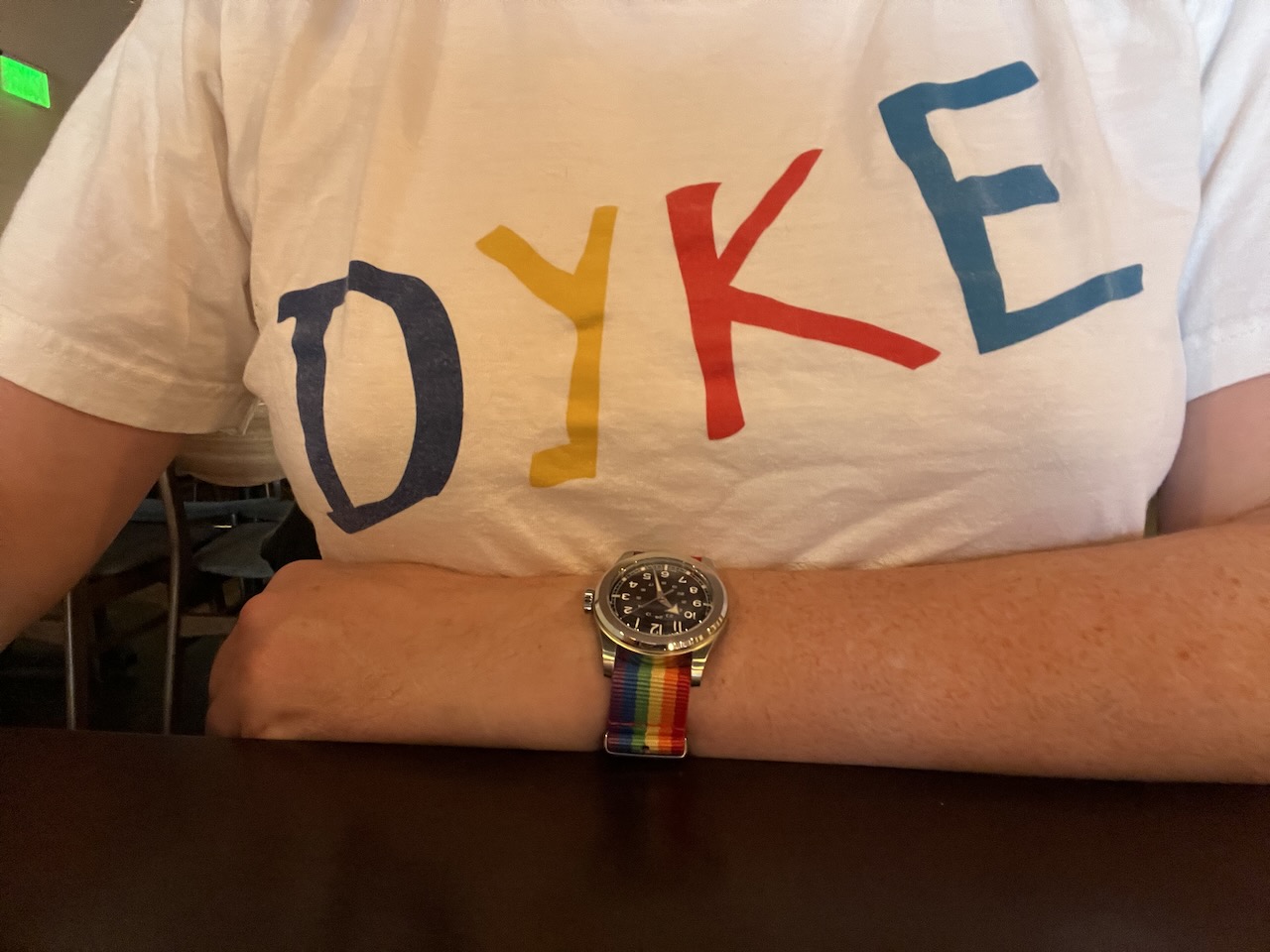 the chest and left arm of a woman. she's wearing a white t-shirt that reads "DYKE" in large, colorful letters across the chest. On her left wrist is a watch on a nylon strap in the colors of the pride flag. The watch (a Serica 4512) has a simple black dial and a broad-arrow hour hand.