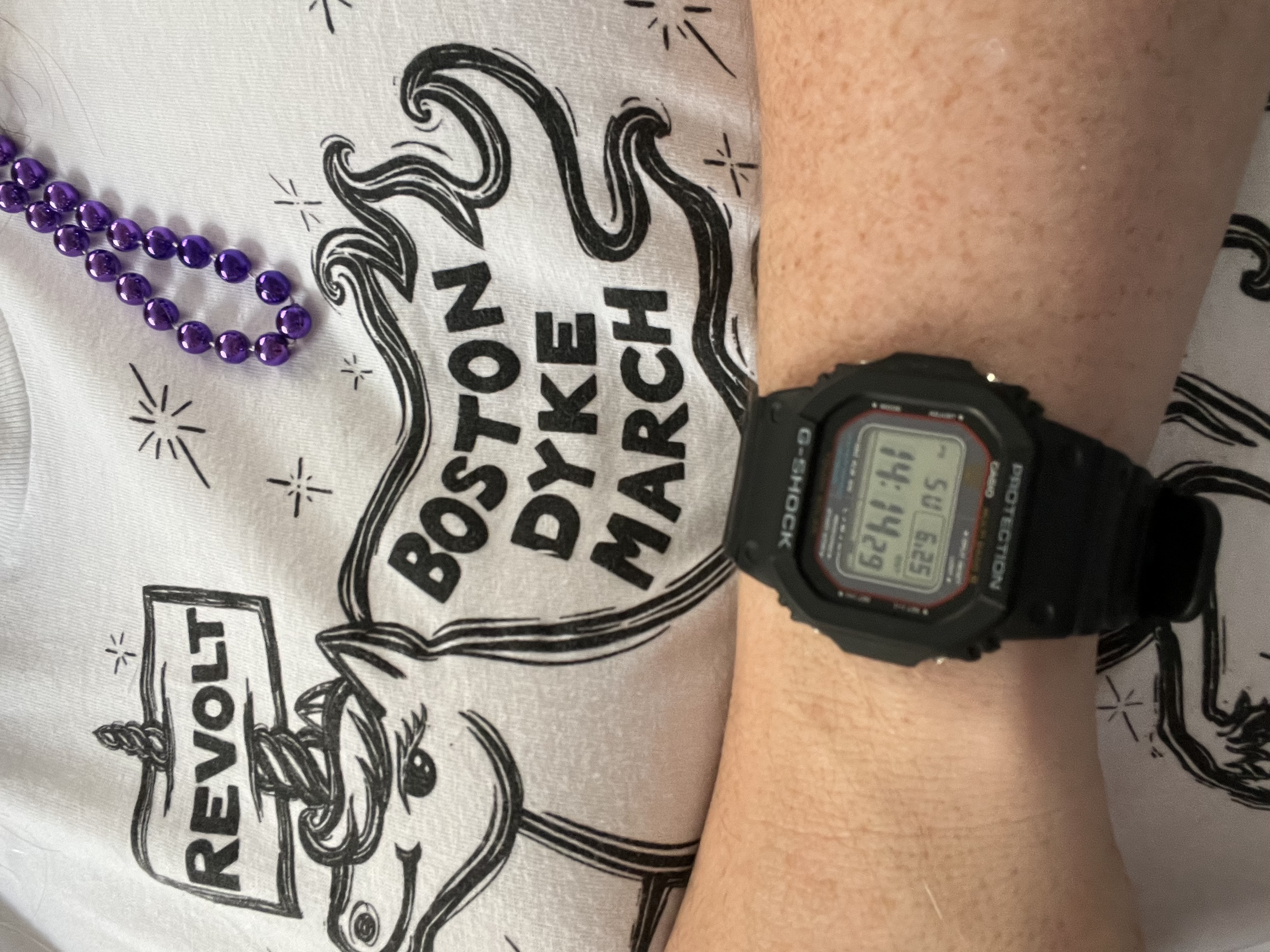 A woman’s chest and left arm. She’s wearing a shirt with a unicorn on it. In the mane of the unicorn are the words BOSTON DYKE MARCH. I paled on the unicorn’s horn is a sign reading REVOLT.On the woman’s left wrist is a chunky black digital watch.