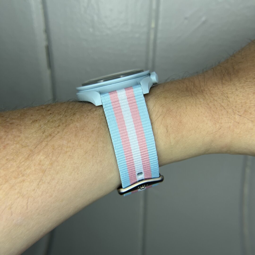 A human wrist from the side with a wrist watch on. The watch can't be seen very well, but it's light blue. The strap the watch is on is colored blue, pink, and white, in the pattern of the transgender pride flag.