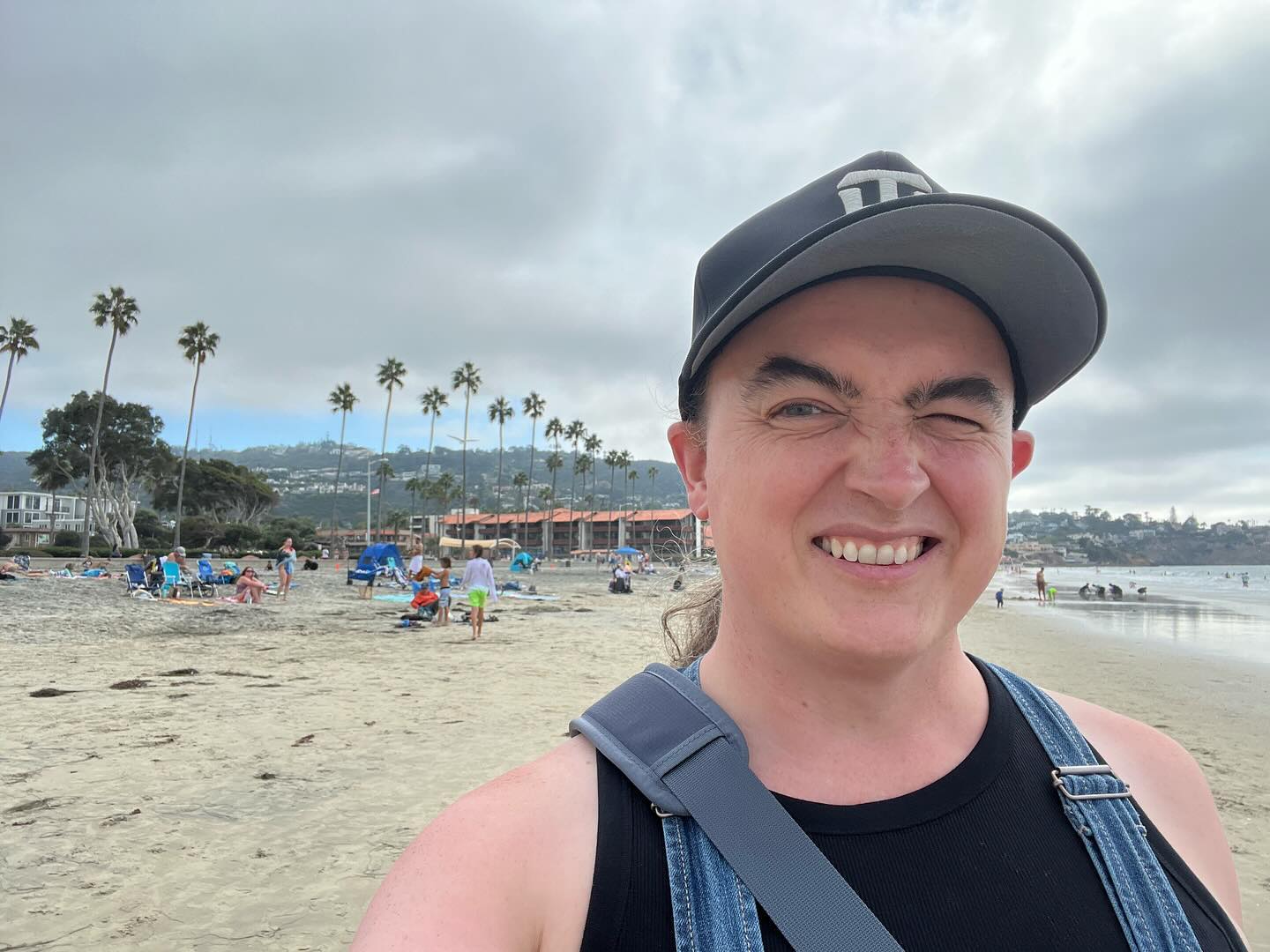 A smiling selfie of me.
I’m standing on the beach at La Jolla Shores; there is a line of palm trees behind me.
I’m squinting in the sun.