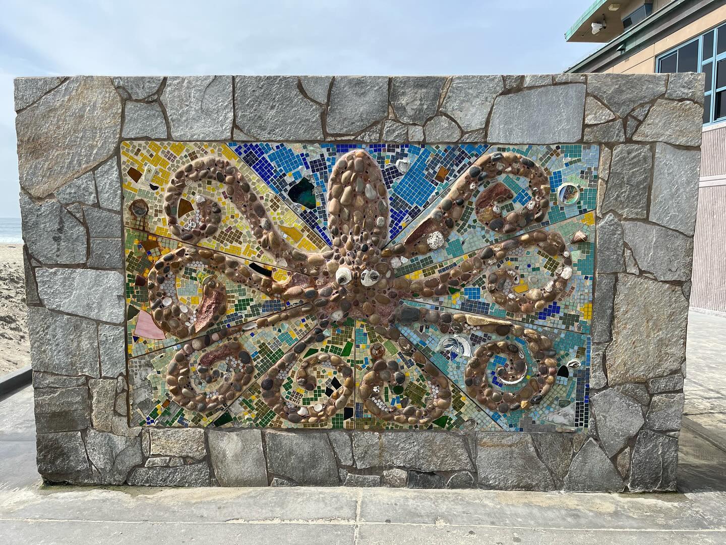 A stone wall near a beach depicts an octopus using stone and title.