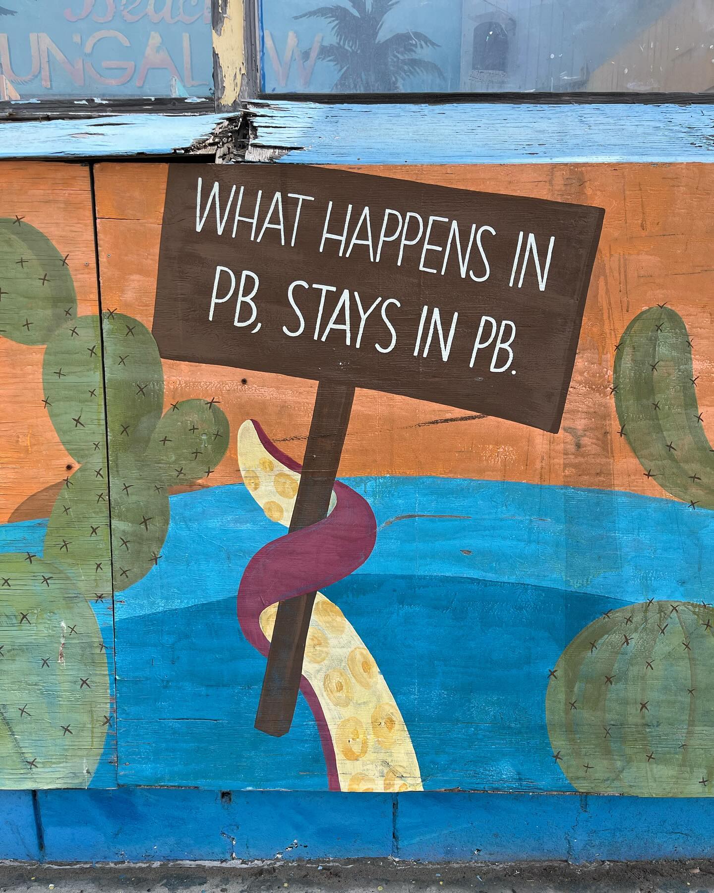 A painting depicts the tentacle of an octopus reaching out from the ocean.
It’s holding a protest sign which reads ‘What happens in PB, stays in PB.’
(PB is Pacific Beach, a neighborhood of San Diego, California.)