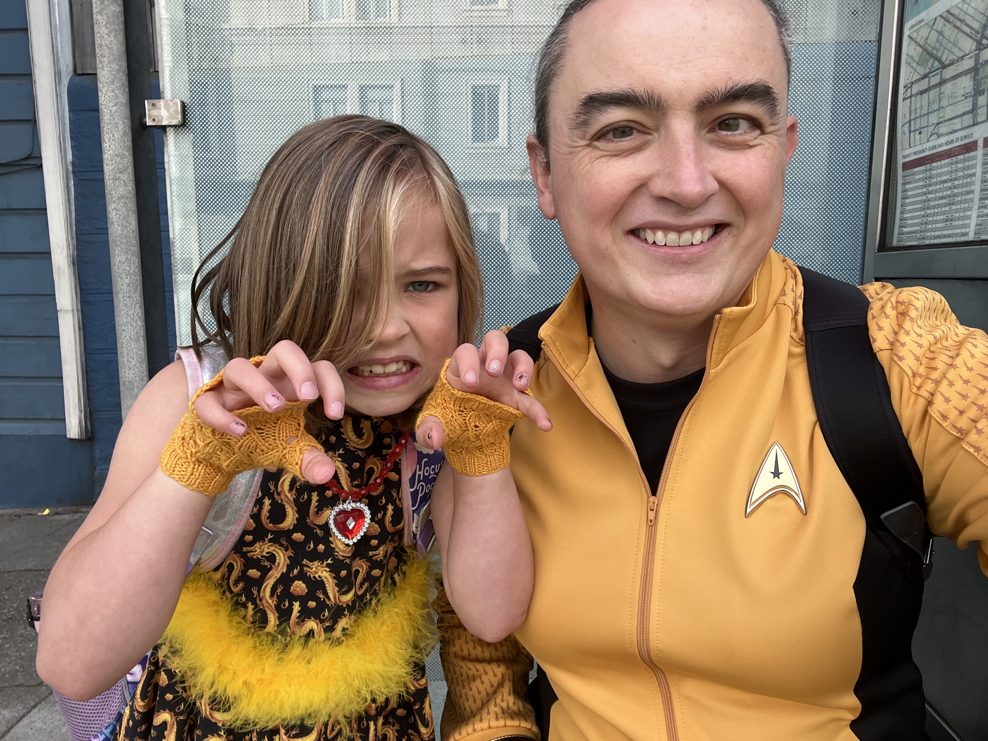 A selfie of EJ and I sitting at a bus stop.
She’s dressed as a golden dragon, with scaly hand-knit fingerless gloves and a homemade black dress with a gold-colored Chinese-style dragon pattern on it.
I’m wearing a command division Star Trek uniform from Strange New Worlds.
