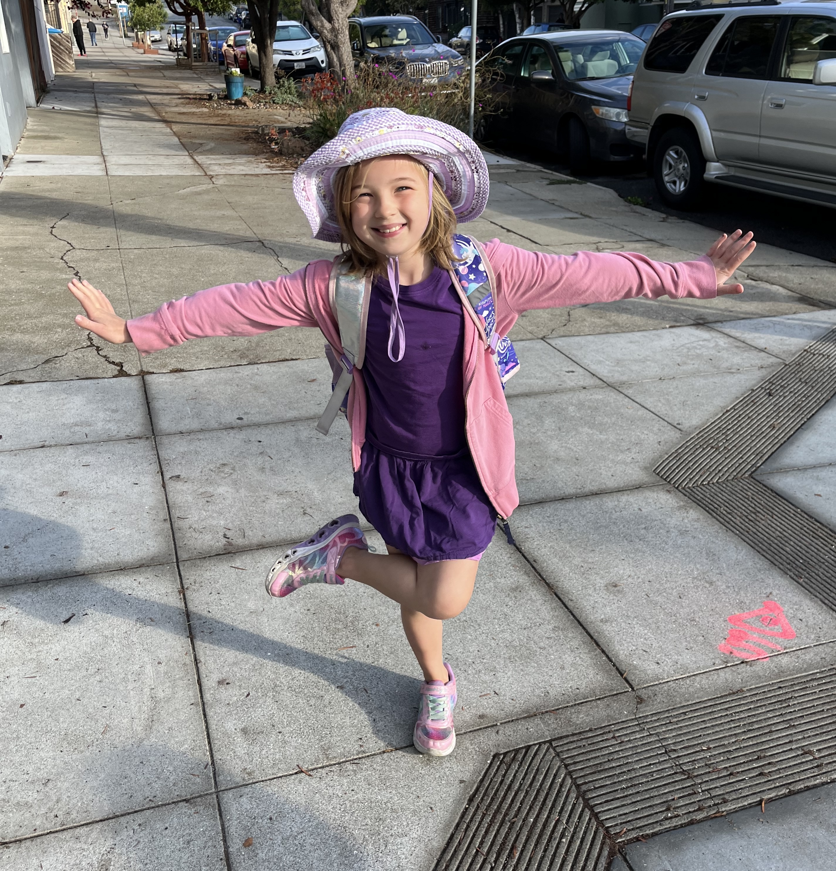 EJ stands on one leg with her arms sticking out.
She’s smiling and squinting. She’s wearing a purple T-shirt with a matching purple skirt, a pink hoodie, a lavendar and white hat, and pink shoes.
The straps of a backpack are visible.