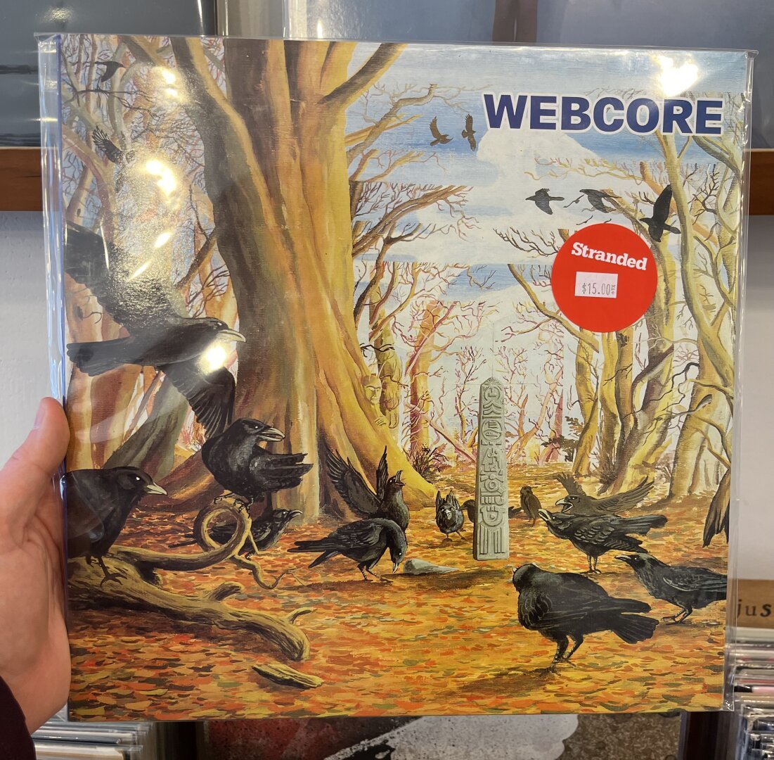 Someone is holding up a record.

The cover depicts a murder of crows in a forest. None of the trees have leaves; the forest floor is covered with them. In the middle of the forest stands a stone obelisk with hieroglyphics on it.

The only word that appears on the cover is "WEBCORE." There's a sticker that denotes it's being sold at a record store called Stranded. The price is $15.00.