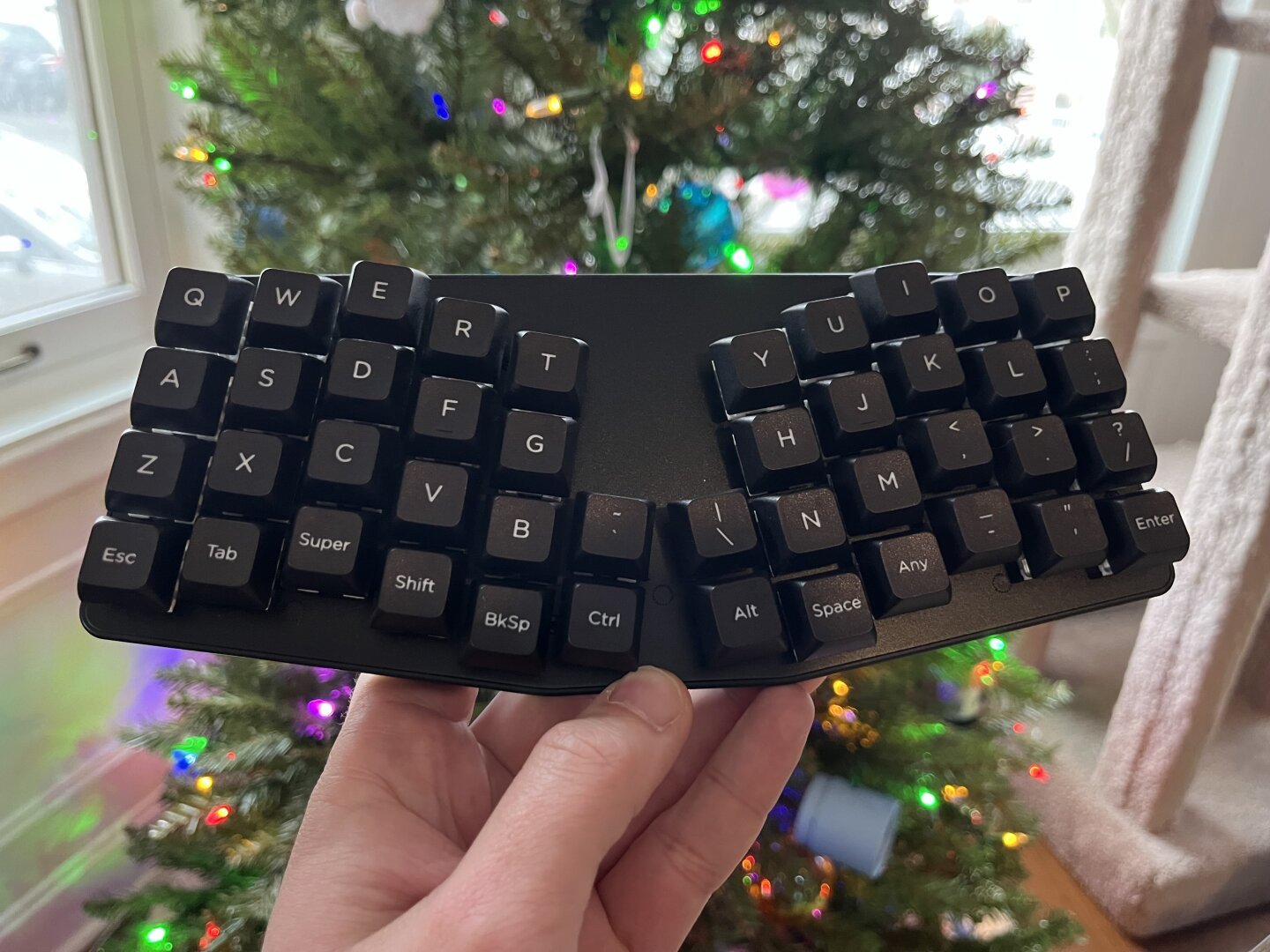 A small keyboard is being held in front of a lit Christmas tree. The keyboard has an unusual, split layout.
