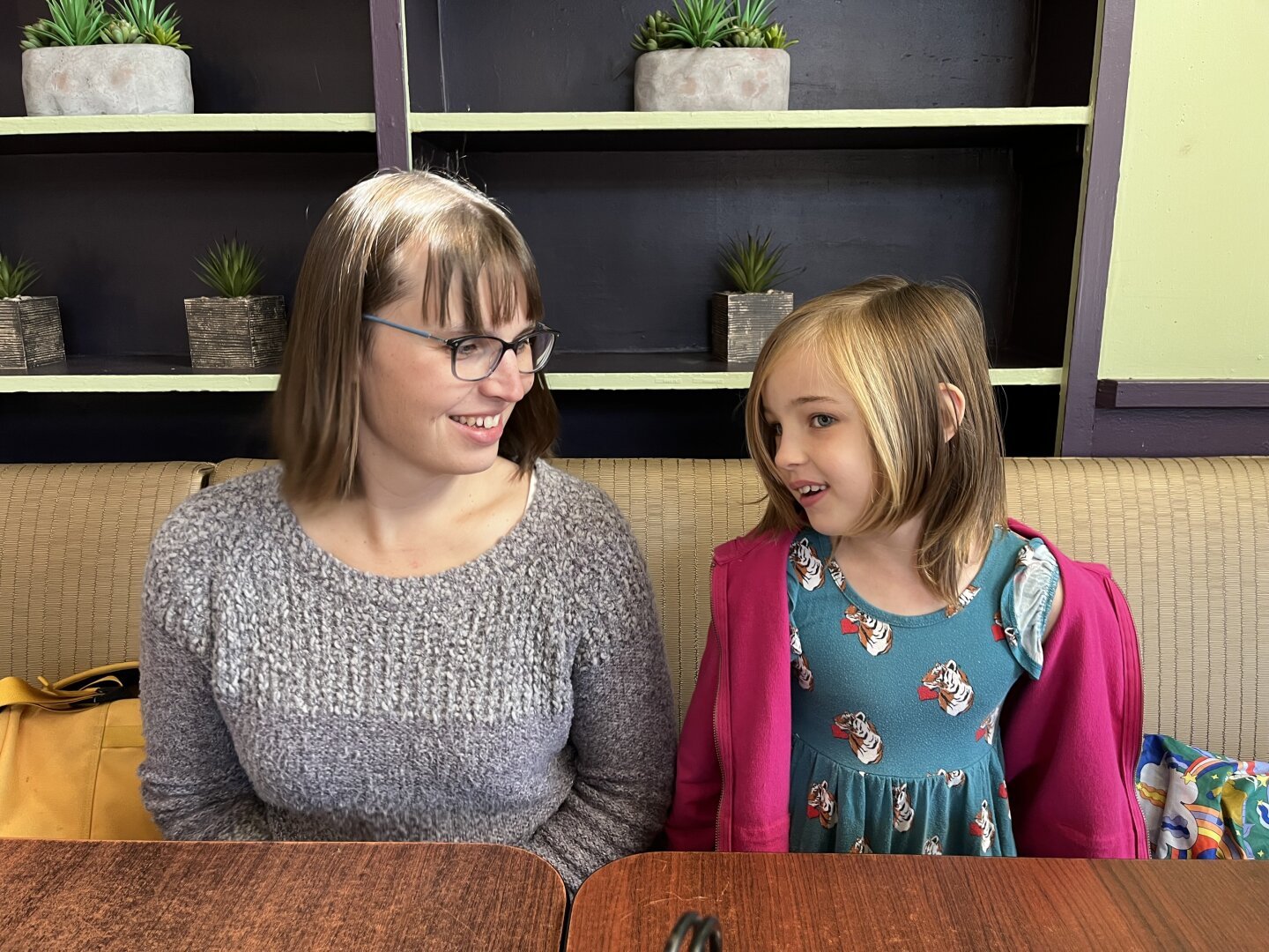 Eryn and EJ sit on one side of a table at a restaurant. Eryn is wearing a very cozy gray sweater. EJ has a pink hoodie on over a green dress. Eryn is looking at EJ and smiling. EJ is smiling and side-eyeing the camera.