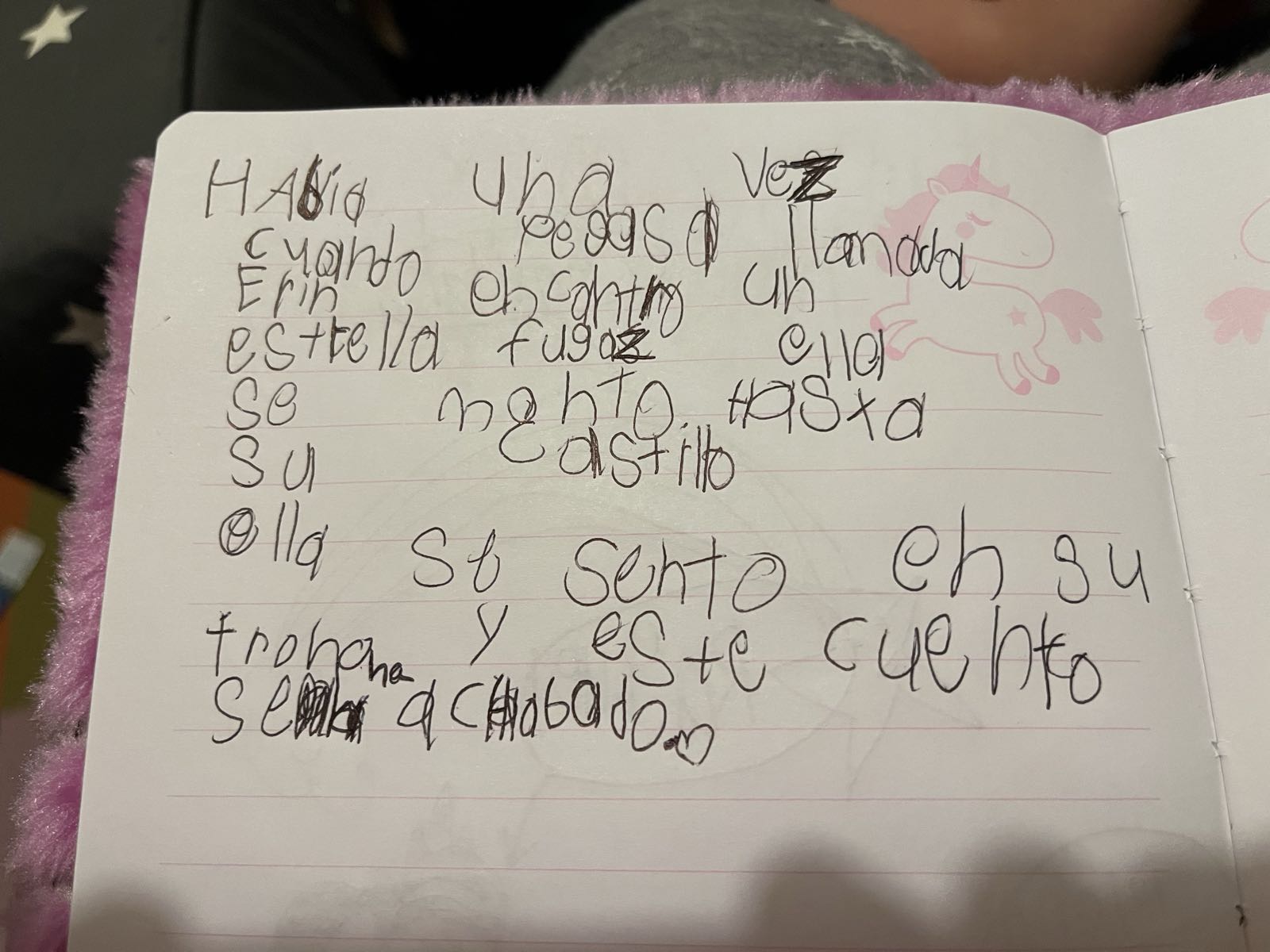 A fuzzy pink notebook is open on a table.
A Spanish-language story is written out in a child’s handwriting.
The story reads: “Había una vez cuando [una] pegasa llamada Erin encontró una estrella fugaz. Ella se montó hasta su castillo. Ella se sentó en su trono y este cuento se ha acabado. 🩷”