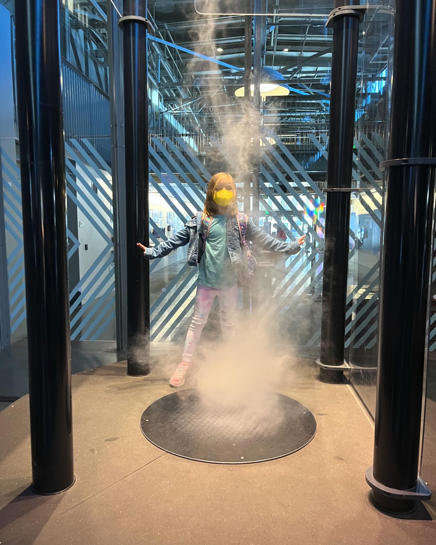 A girl stands with her legs apart and arms spread above a grate.
What appears to be steam is coming through the grate.
(It is not hot and the child is not in pain.)
She’s wearing a blue shirt, tie-dyed pants, a jean jacket, and a yellow face mask.