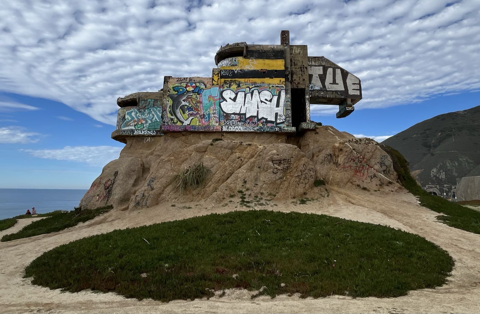 An abandoned concrete structure sits atop a beige stone outcropping in front of a cloudy sky.
The structure is heavily graffitied.
In front of the outcropping there’s a patch of grass surrounded by rock.
To the left of the outcropping the ocean is visible.