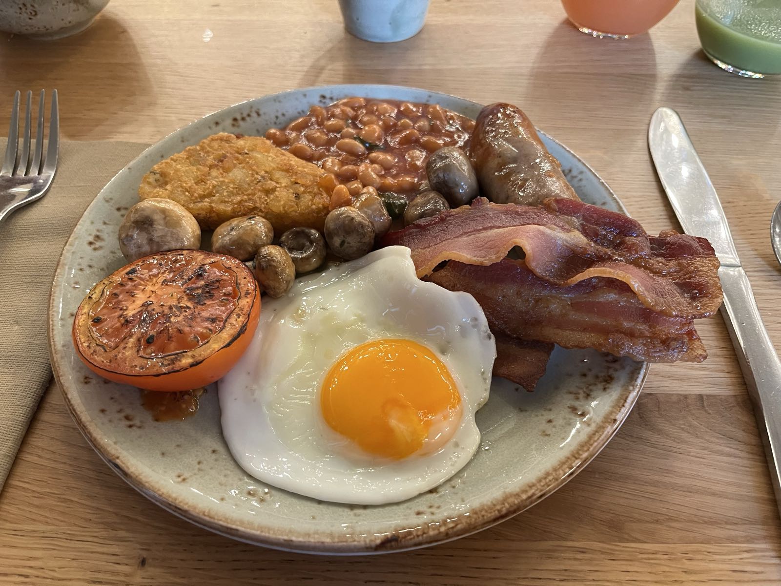 A plate of food.
Clockwise from left, there’s
a grilled tomato,
some mushrooms,
a hash brown,
baked beans,
a banger,
streaky bacon,
and a fried egg.