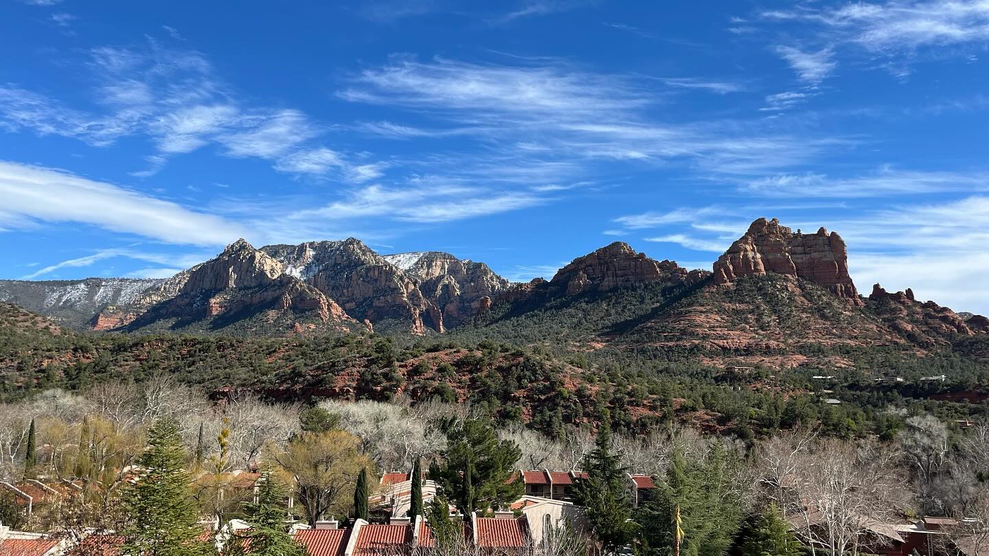 In the distance are a number of hilly desert rock formations. Between here and there, some buildings are nestled among trees.